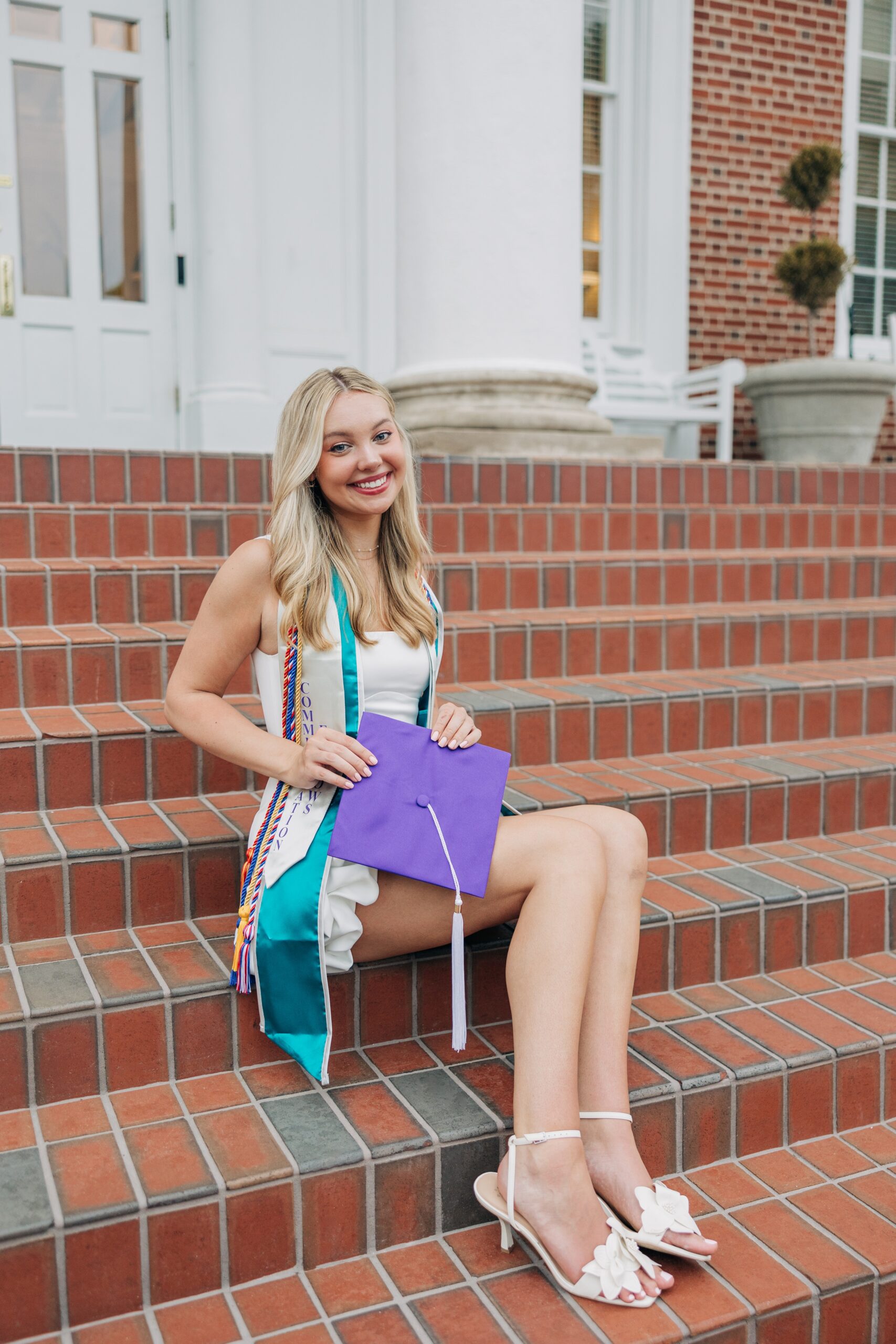 An image from a high point university graduation photographer of a woman sitting on brick stairs in her cords, stole and white dress holding her purple cap