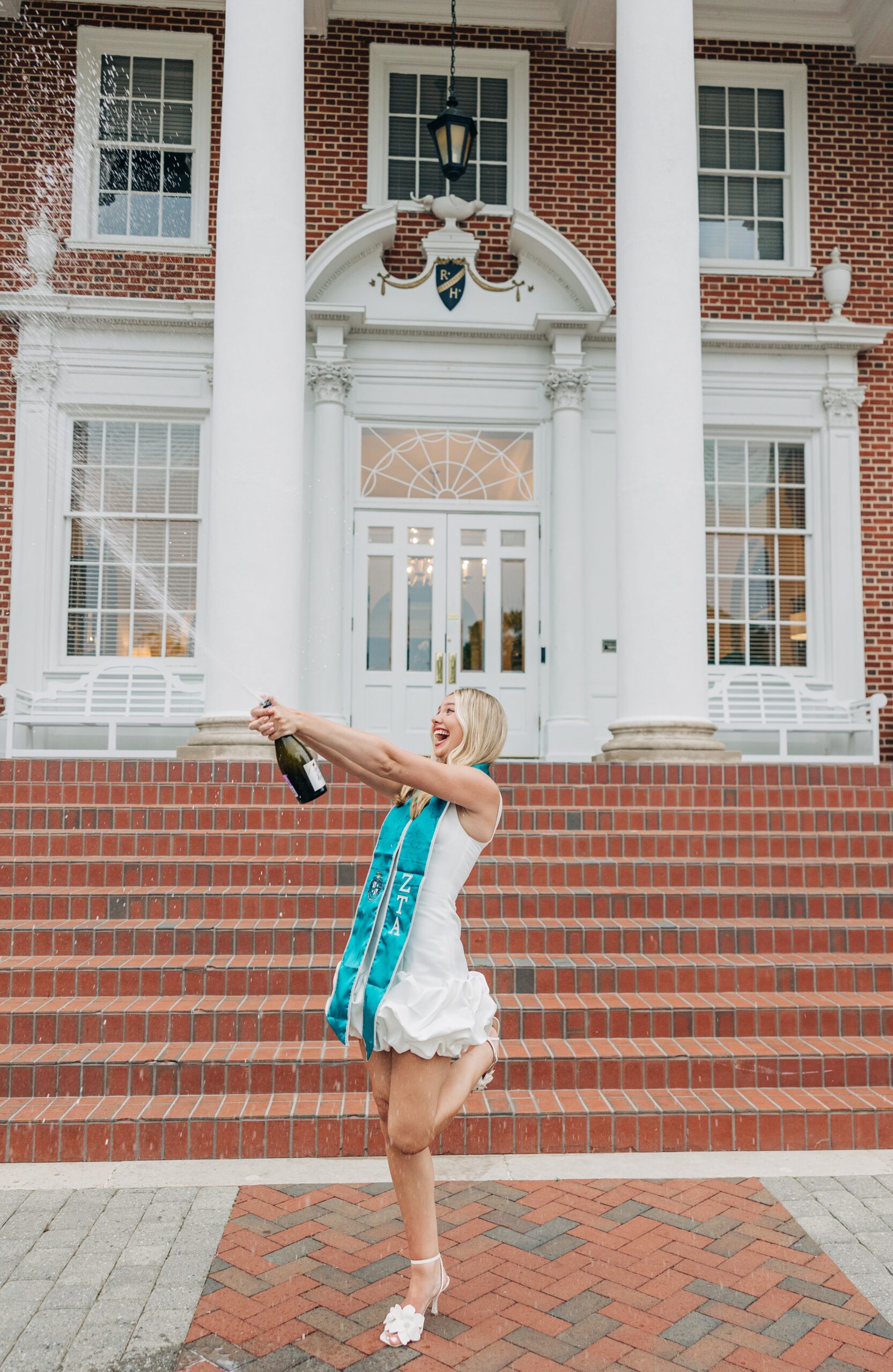 A woman in a white dress pops a bottle of champagne with her sorority stole