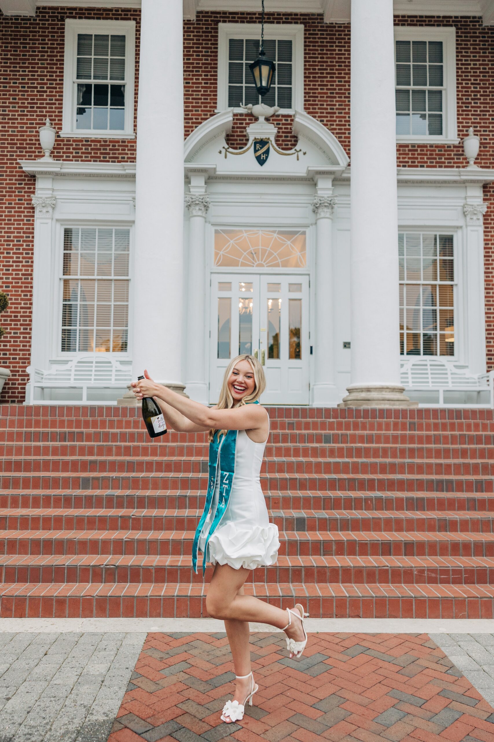 A happy grad pops a bottle of champagne in front of a colonial building steps