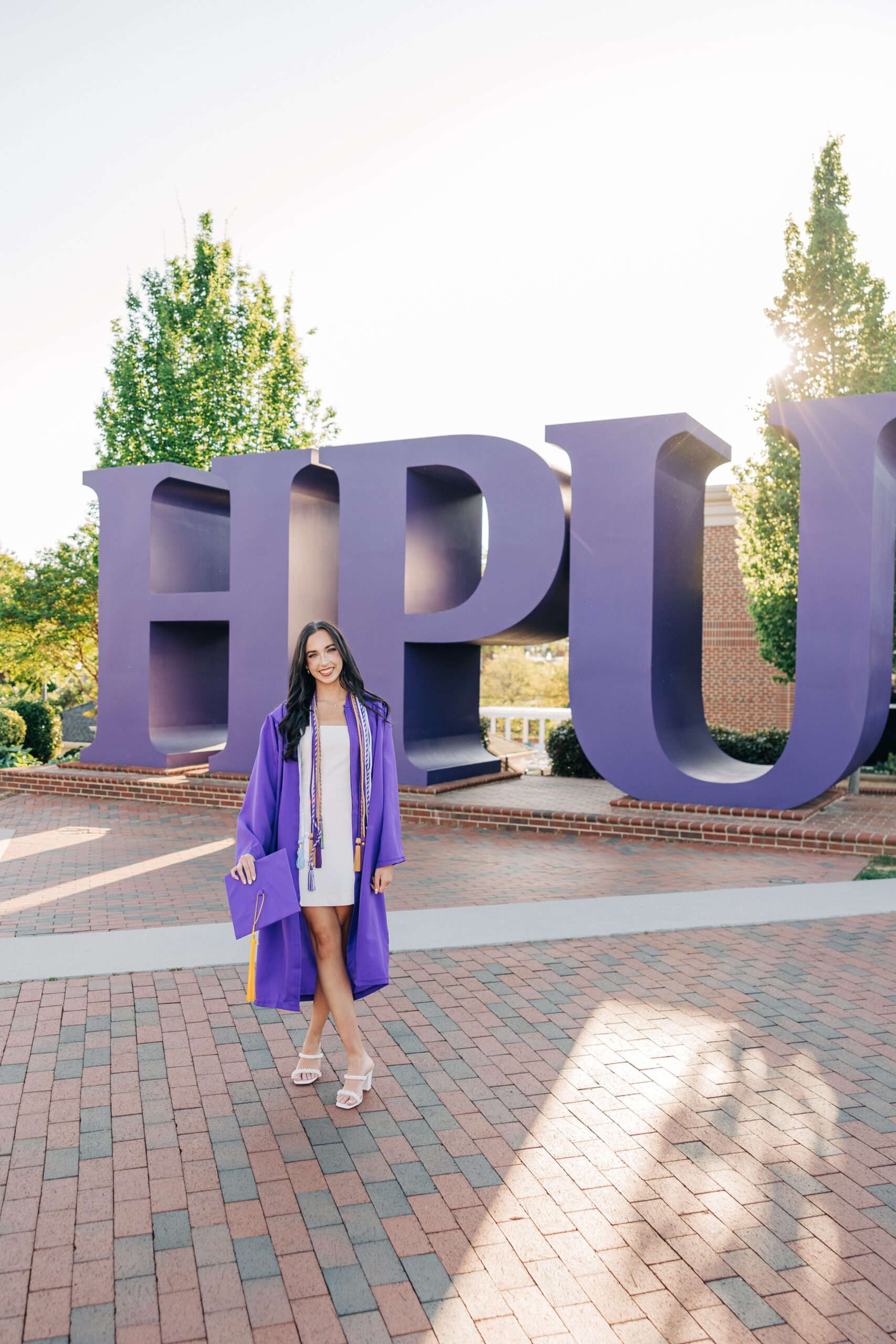 A graduate walks in front of the HPU sign in her cap and gown and white dress