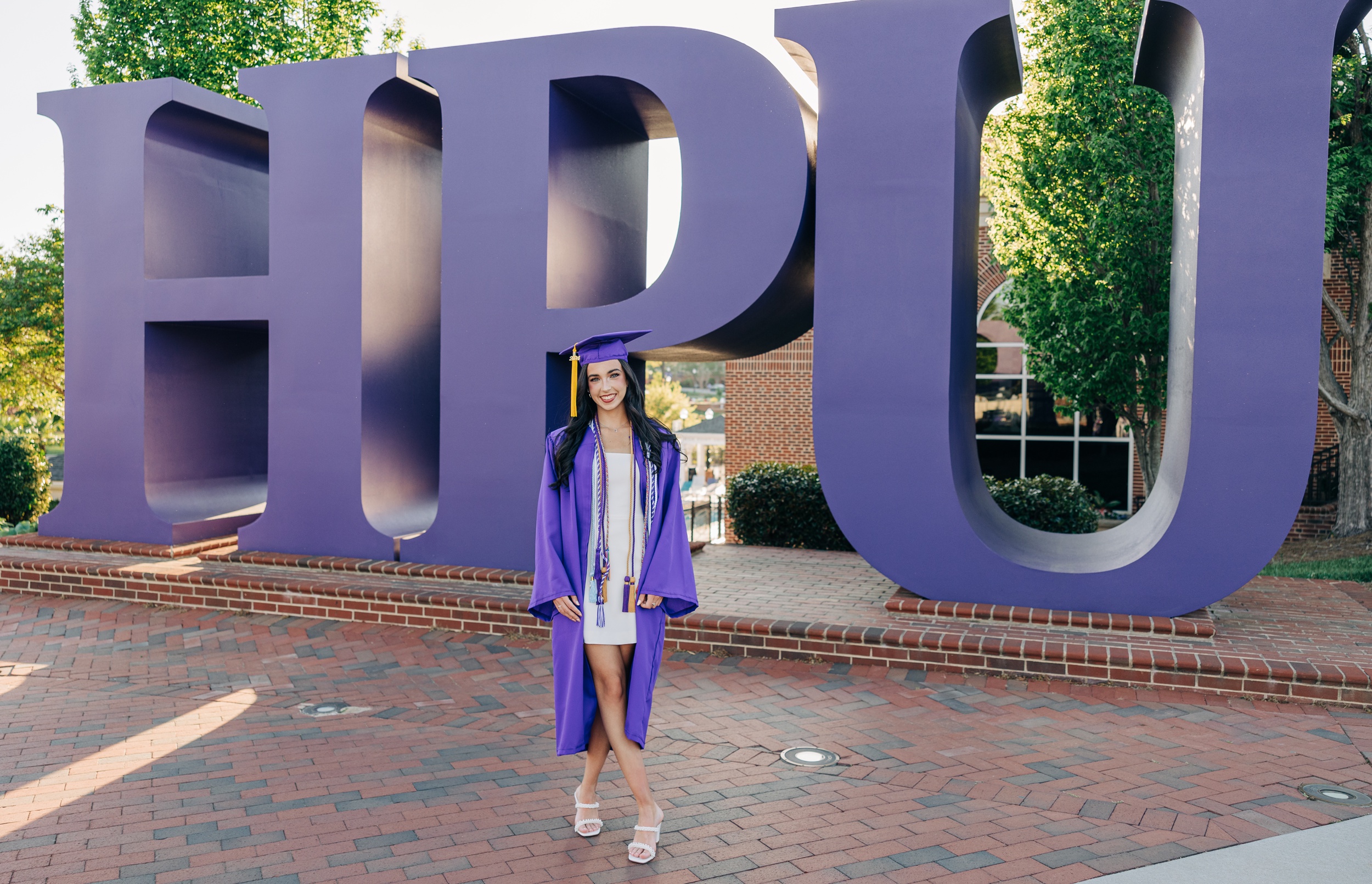 A smiling grad stands in front of the purple sign with her matching cap and gown