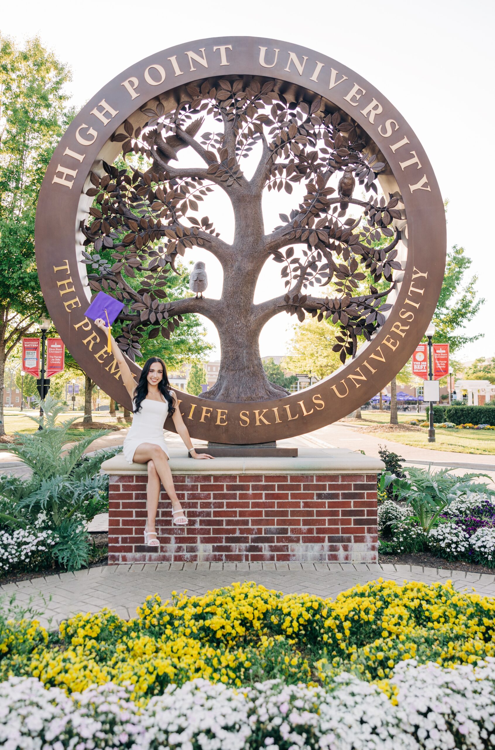 A woman in a white dress celebrates her graduation sitting on the sign with her cap in the air