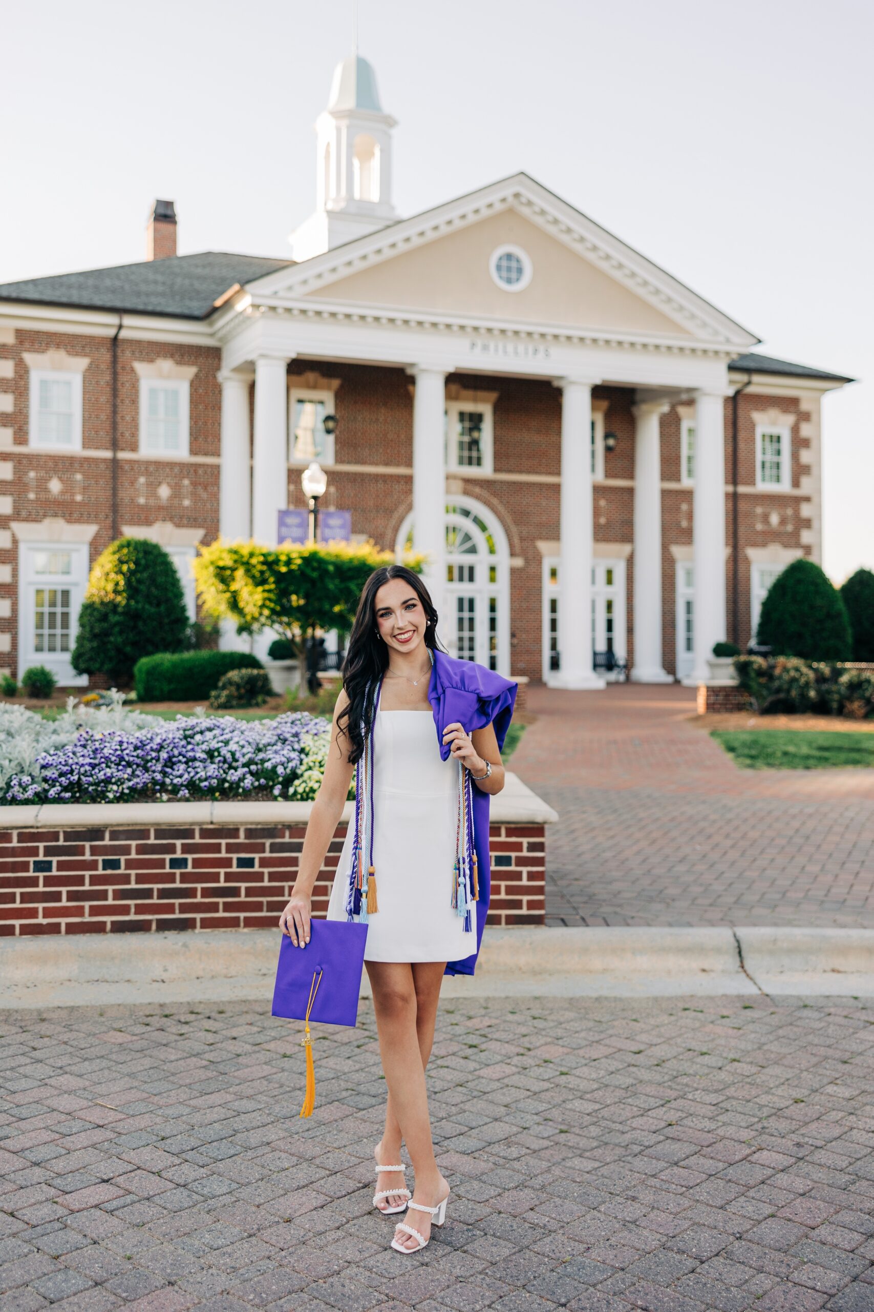 A graduate in a white dress smiles while walking with her gown over her shoulder and cap in her hand