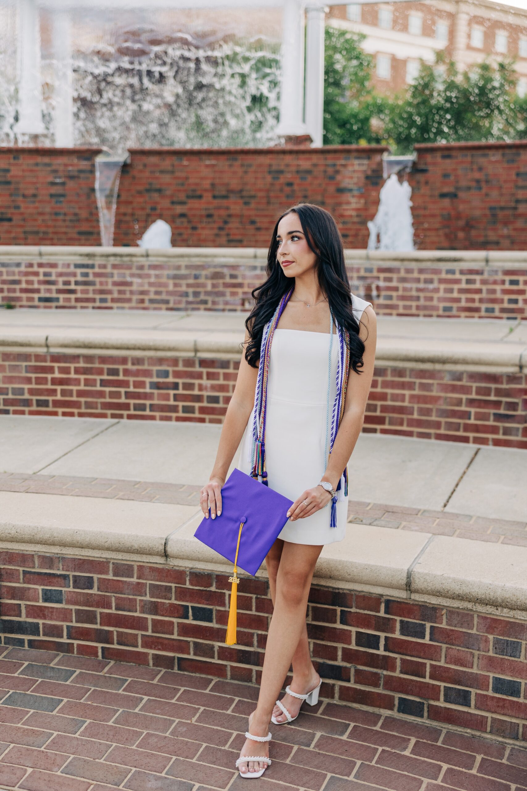 A woman walks in the fountain area holding her purple cap in a white dress