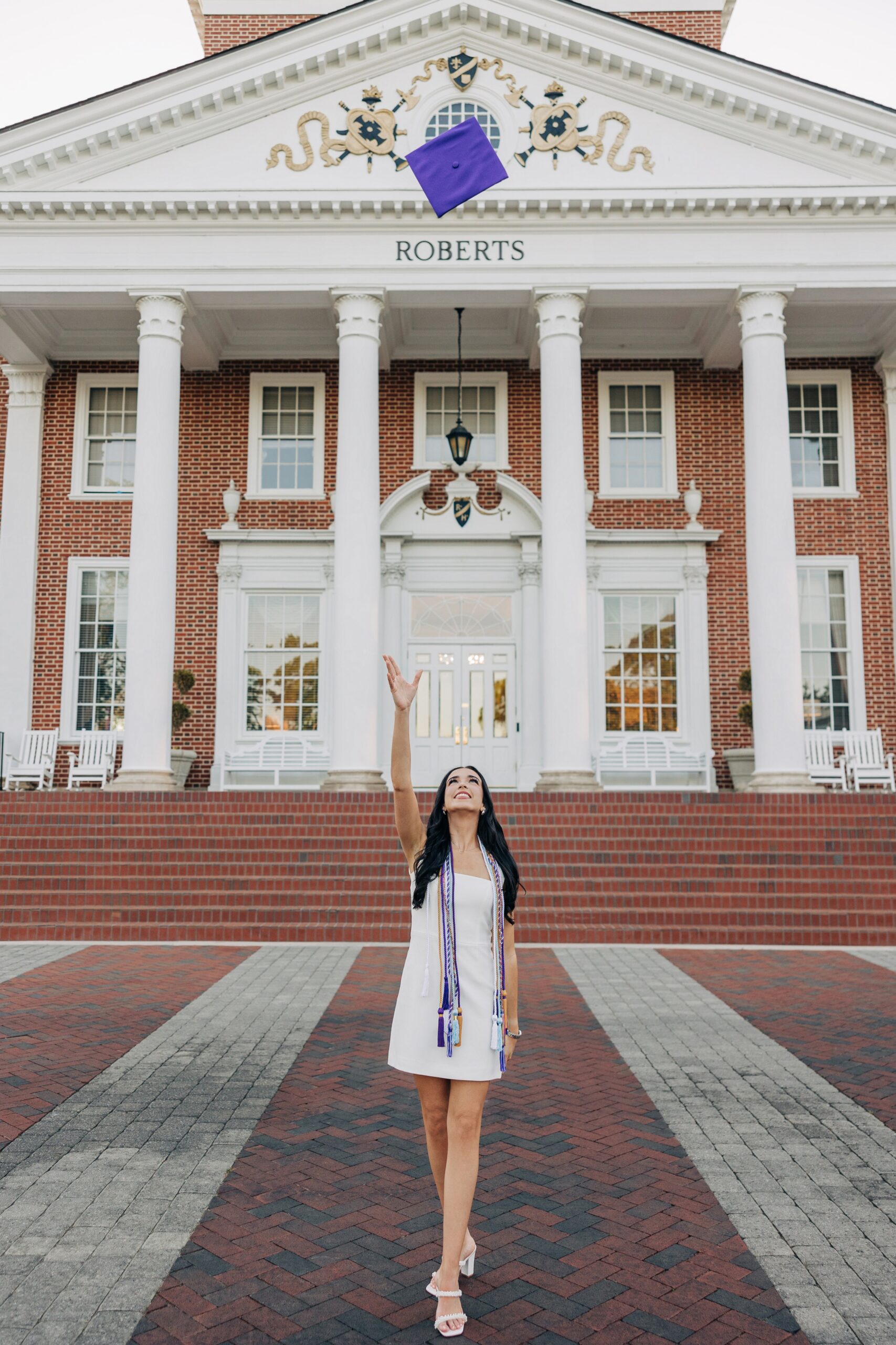 A happy grad tosses her cap while standing on a brick sidewalk in front of the Roberts building captured by a high point university graduation photographer