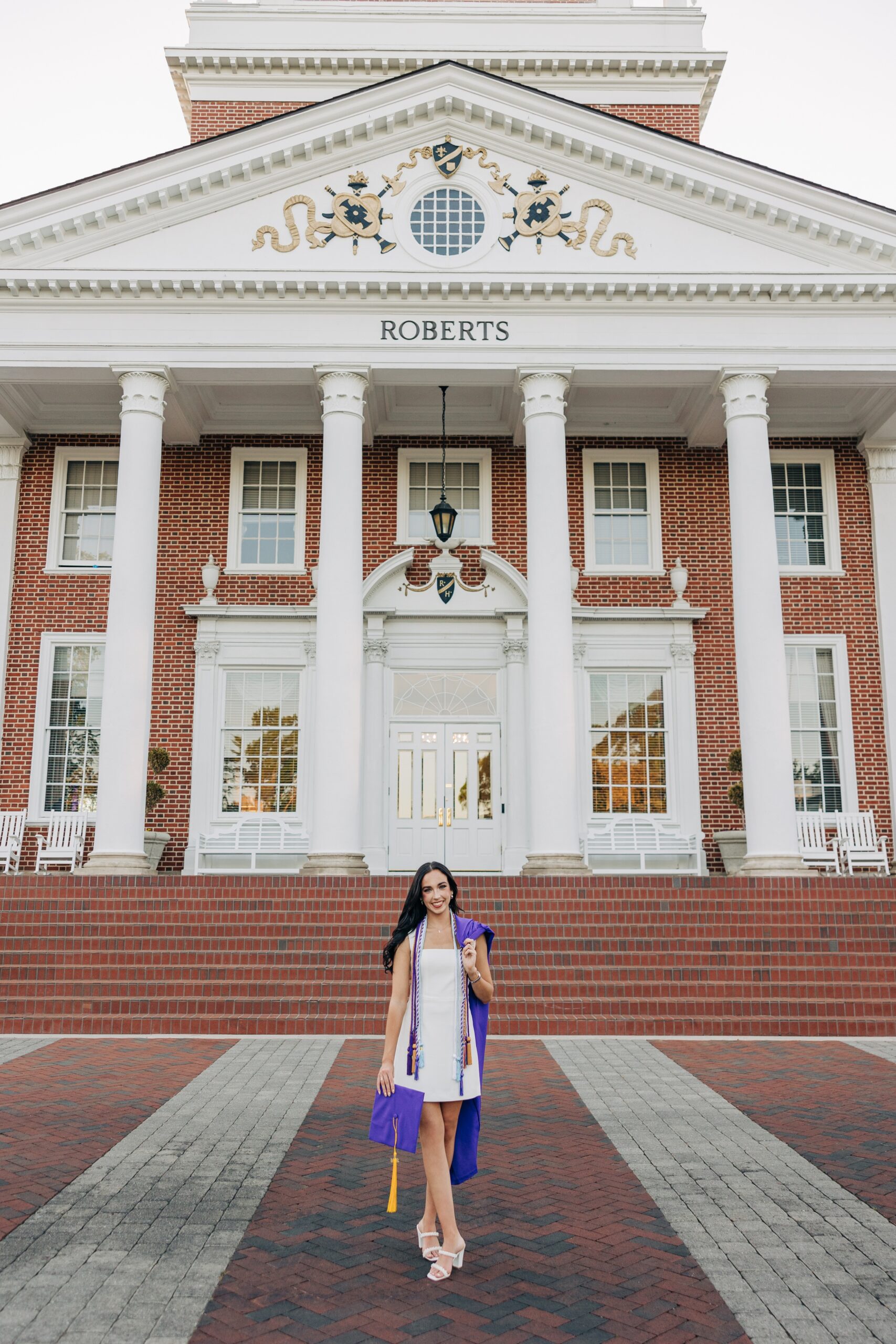 A grad in a white dress wears her chords while standing in front of an old building with her gown over her shoulder for a high point university graduation photographer