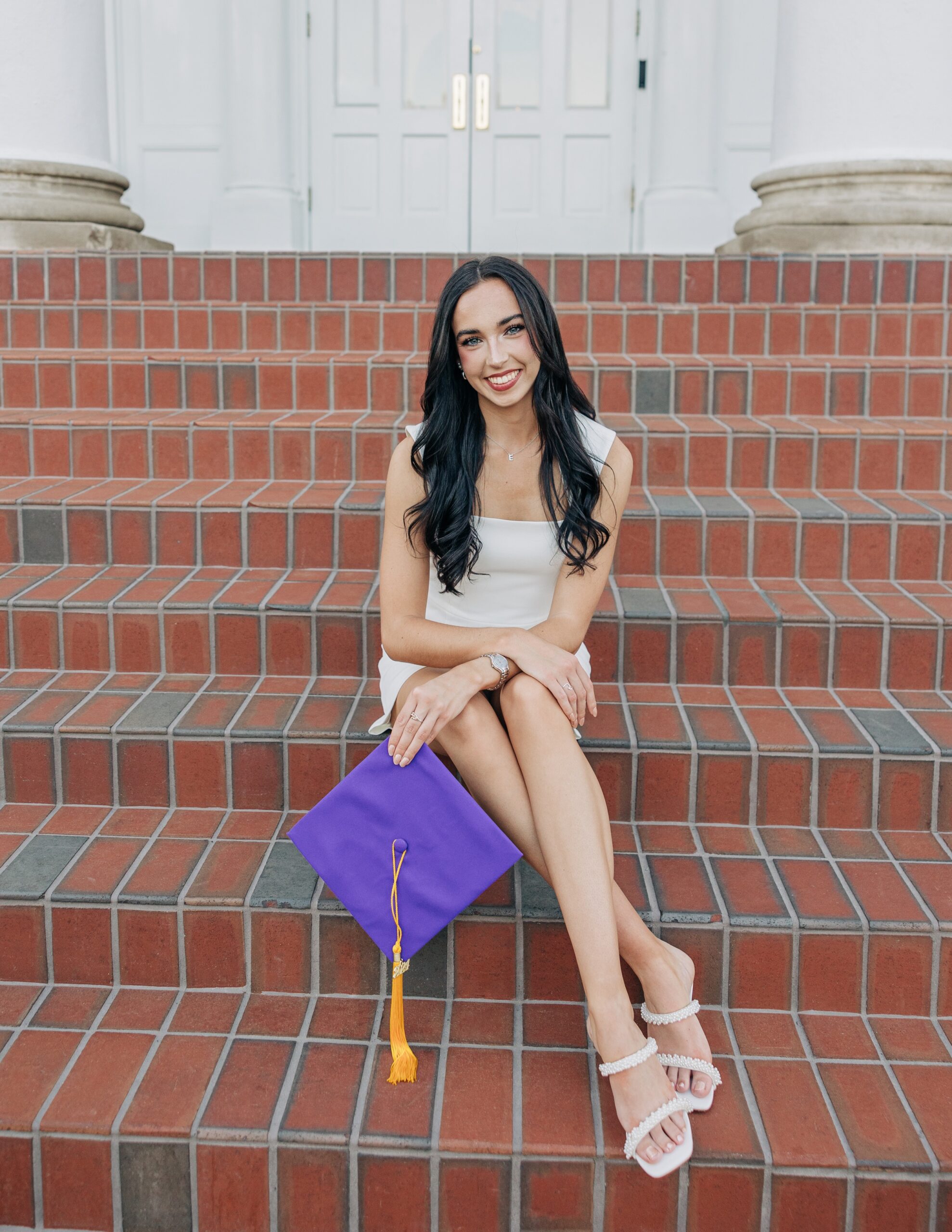 A woman in a white dress sits on brick steps holding her purple cap for her high point university graduation photographer