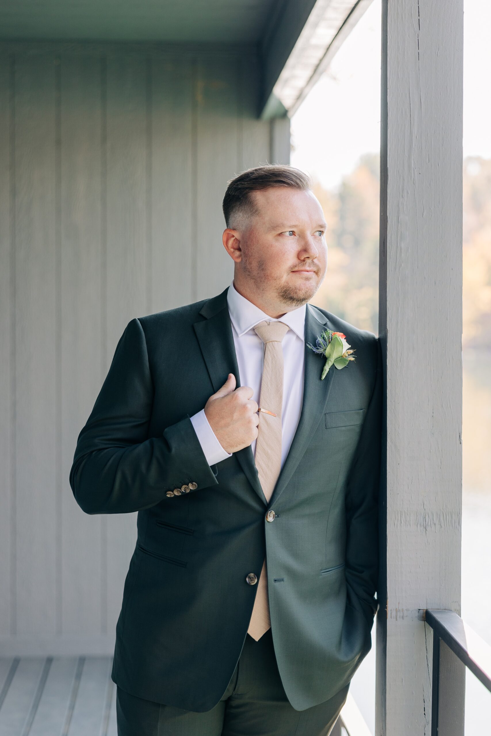 A groom leans on the porch railing holding his green lapel and gazing out to the water at splendor pond flower farm