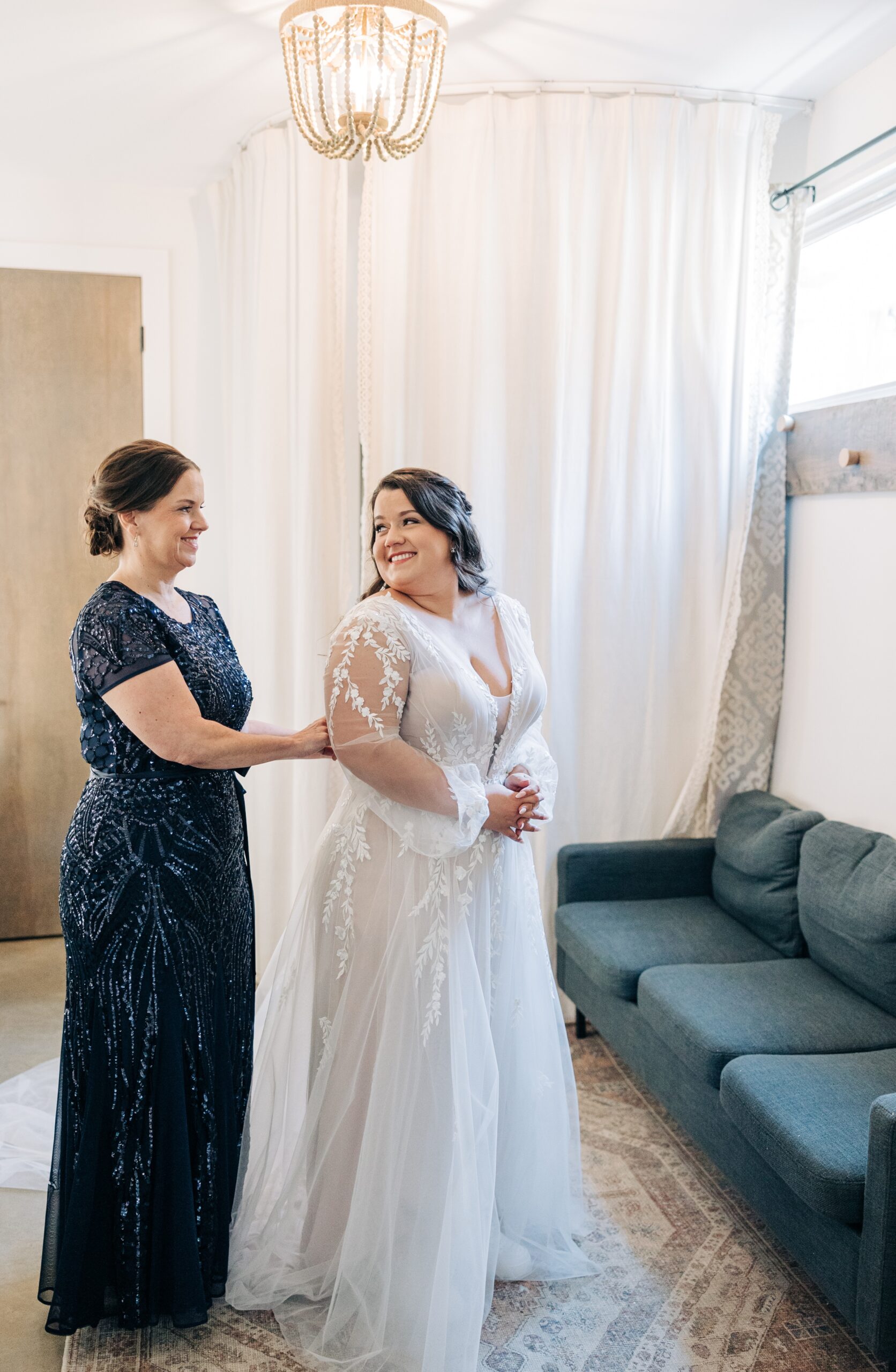 A bride smiles over her shoulder to mom buttoning her into her dress