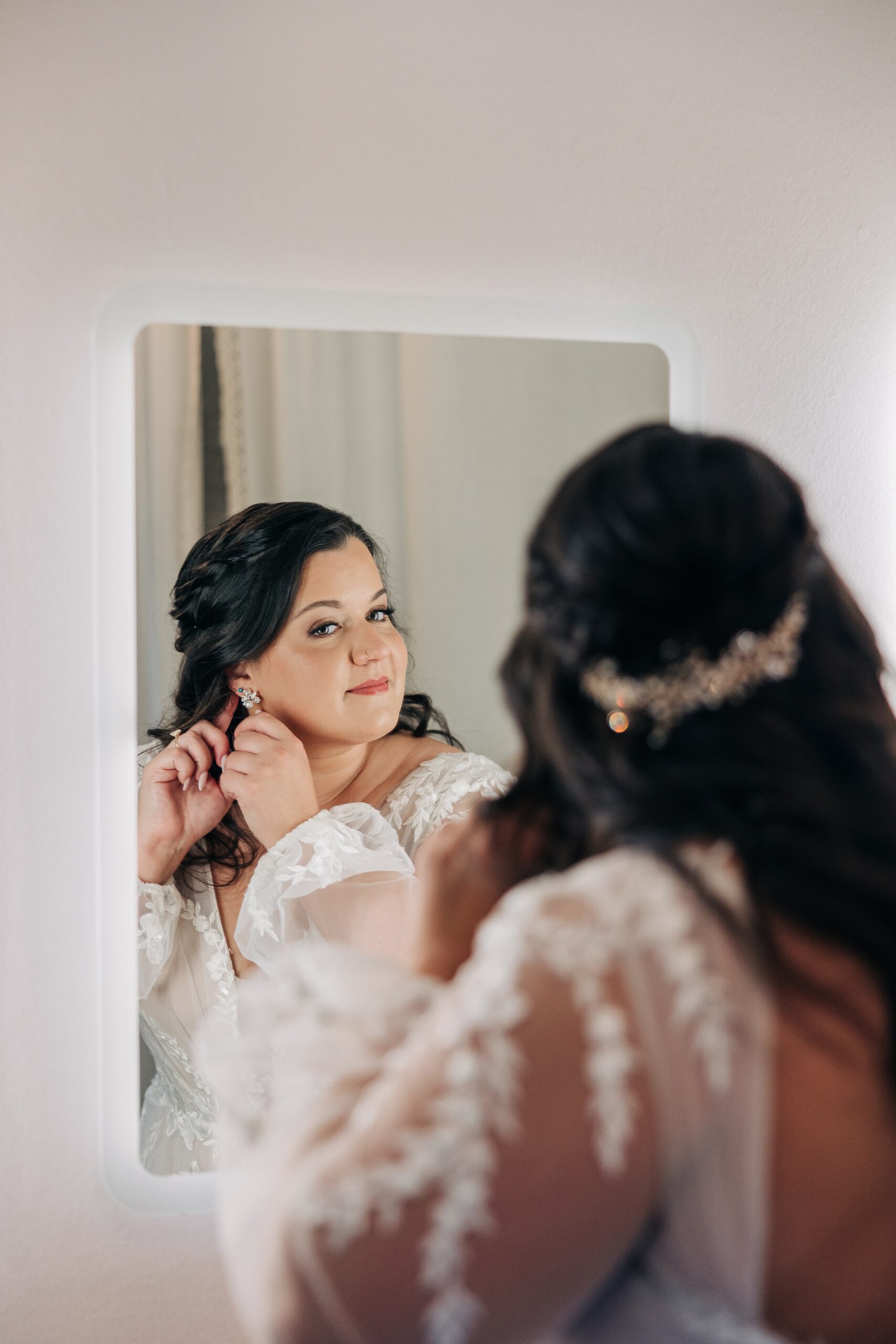 A bride puts on her earring in a mirror in a lace dress