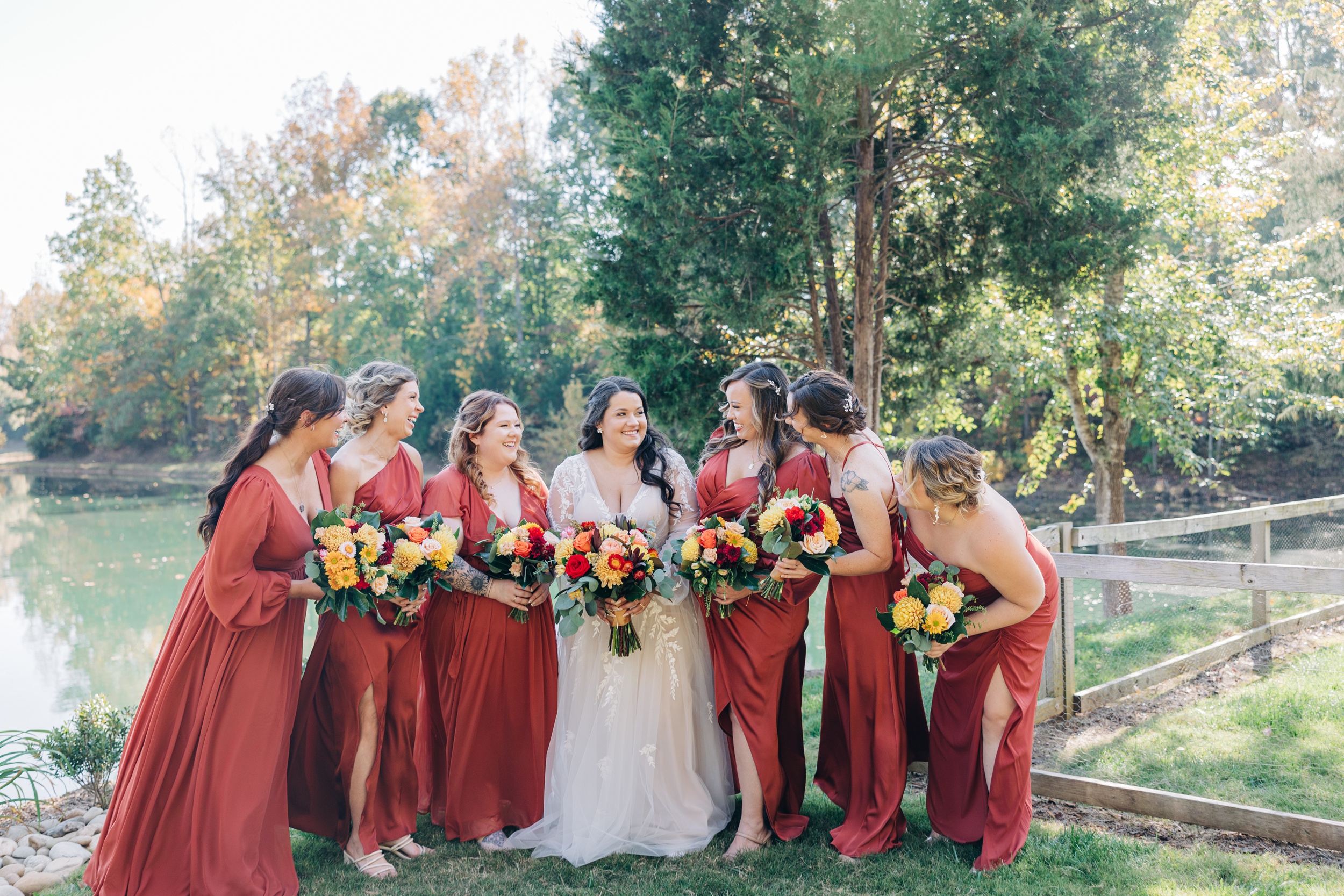 A bride giggles with her bridesmaids in red dresses by the water holding bouquets at splendor pond flower farm