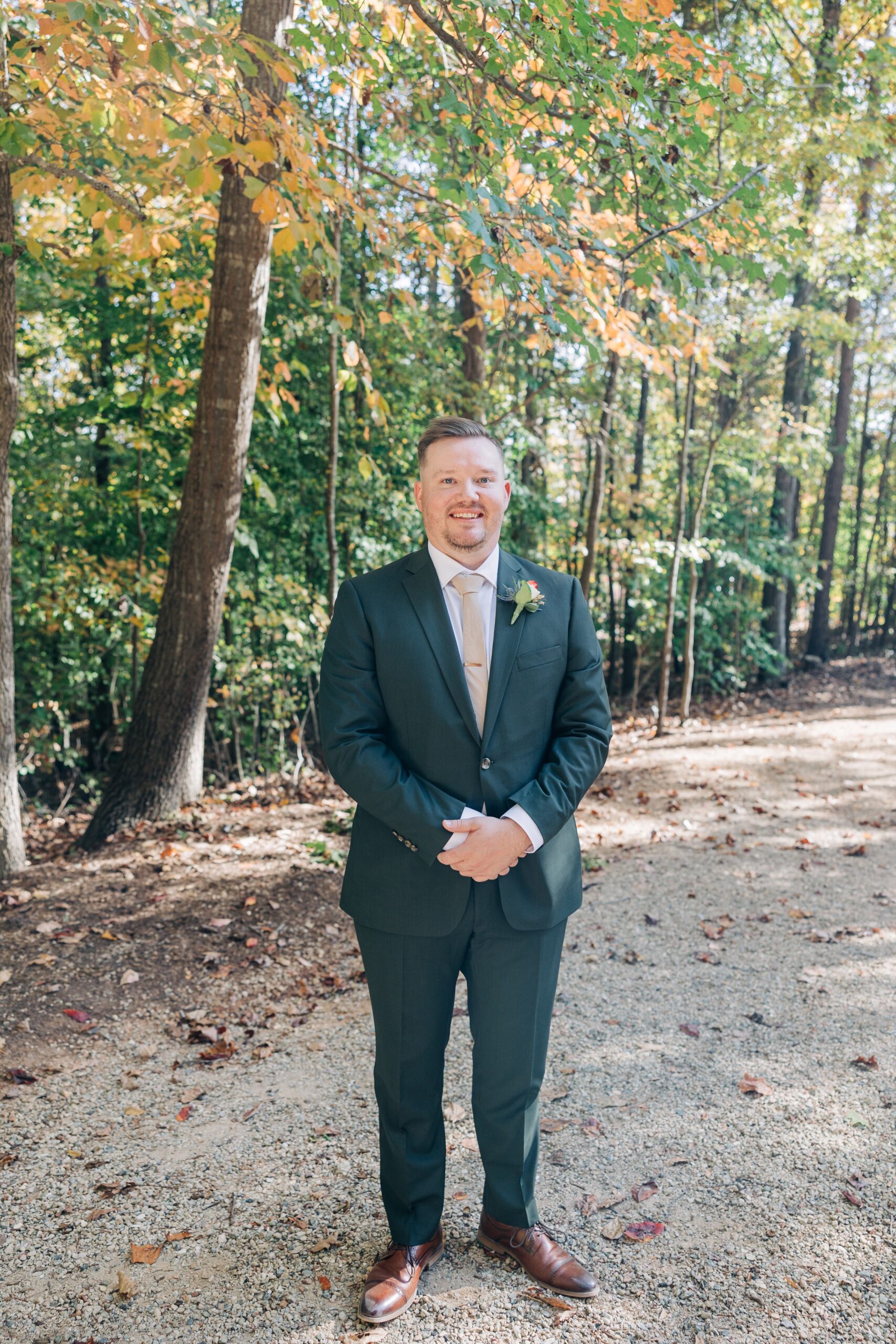 A groom smiles in a green suit in the forest with hands crossed