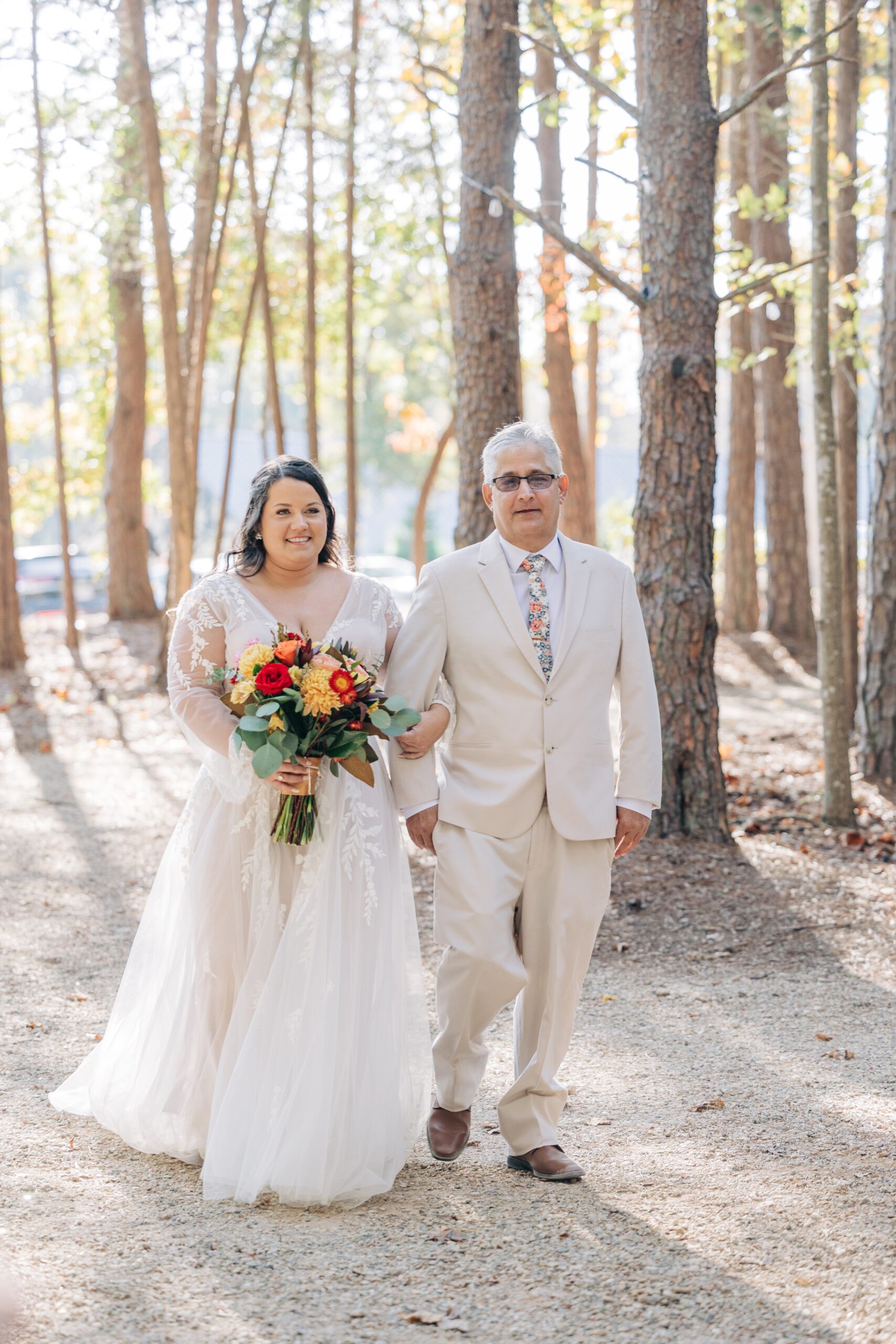 A bride walks down the forest aisle holding her dad's arm in a white suit