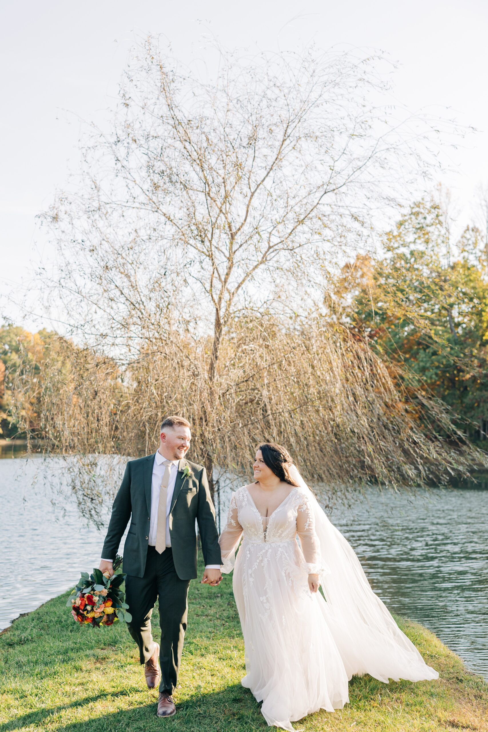 Newlyweds hold hands while walking by a willow tree on the water