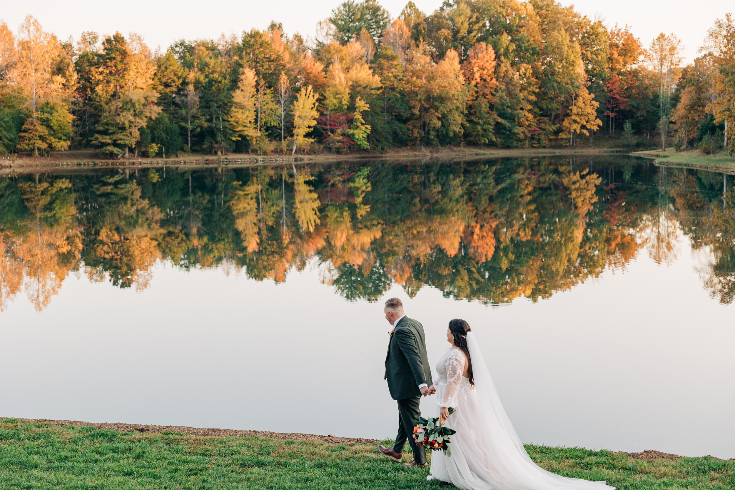 A groom leads his bride by the hand along the water's edge at their splendor pond flower farm wedding