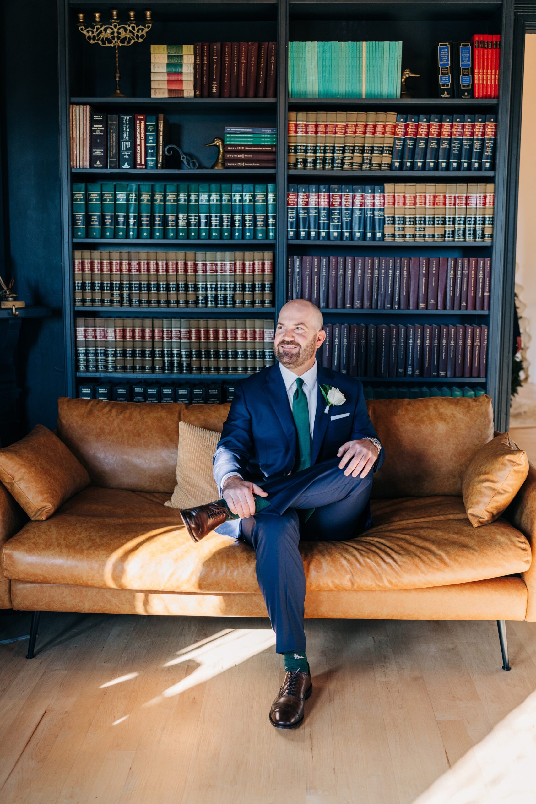 A groom sits in a leather couch under a book care in a blue suit at the crest winston-salem