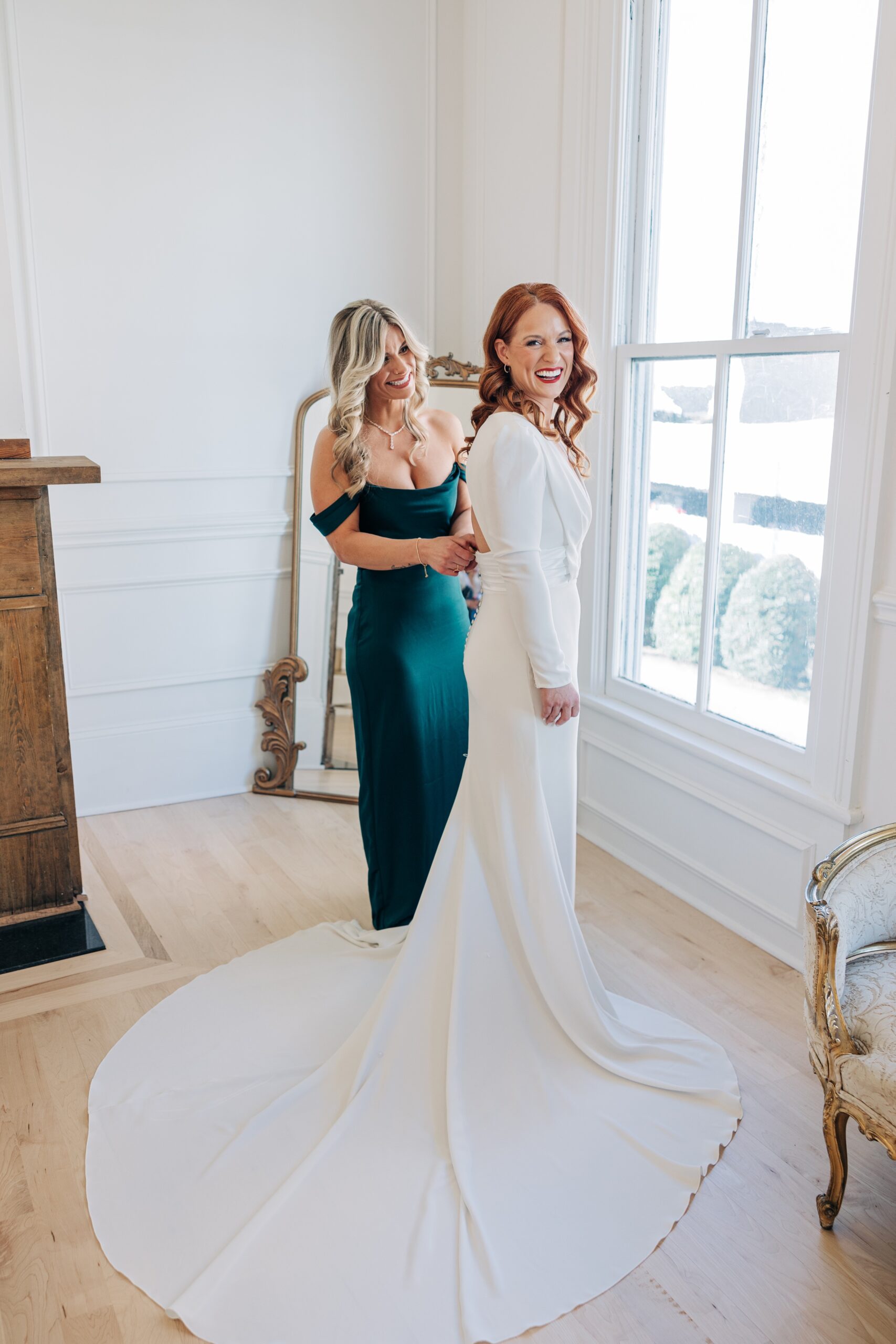 A bridesmaid helps the bride into her dress in a window