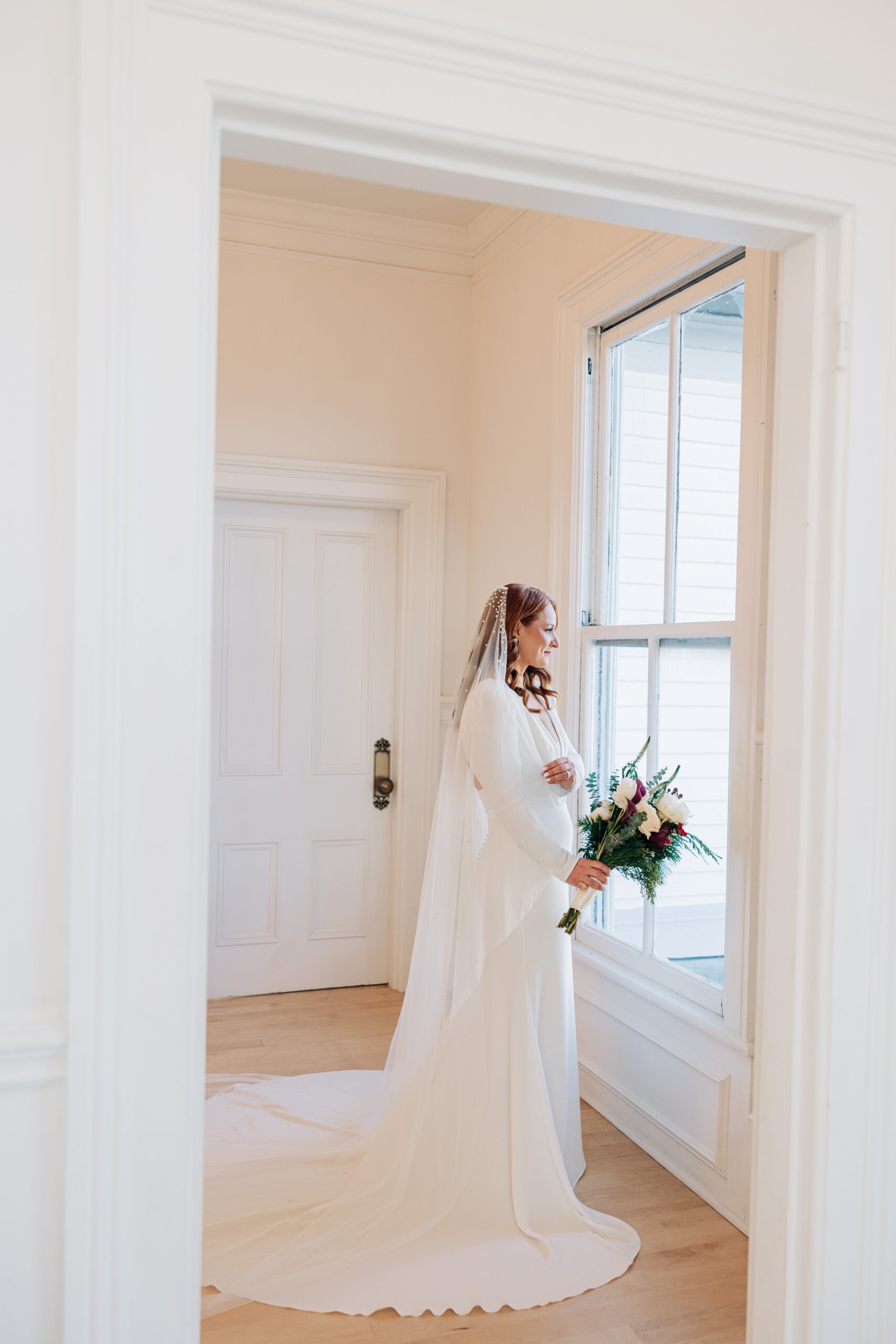 A bride smiles out a window while holding her bouquet