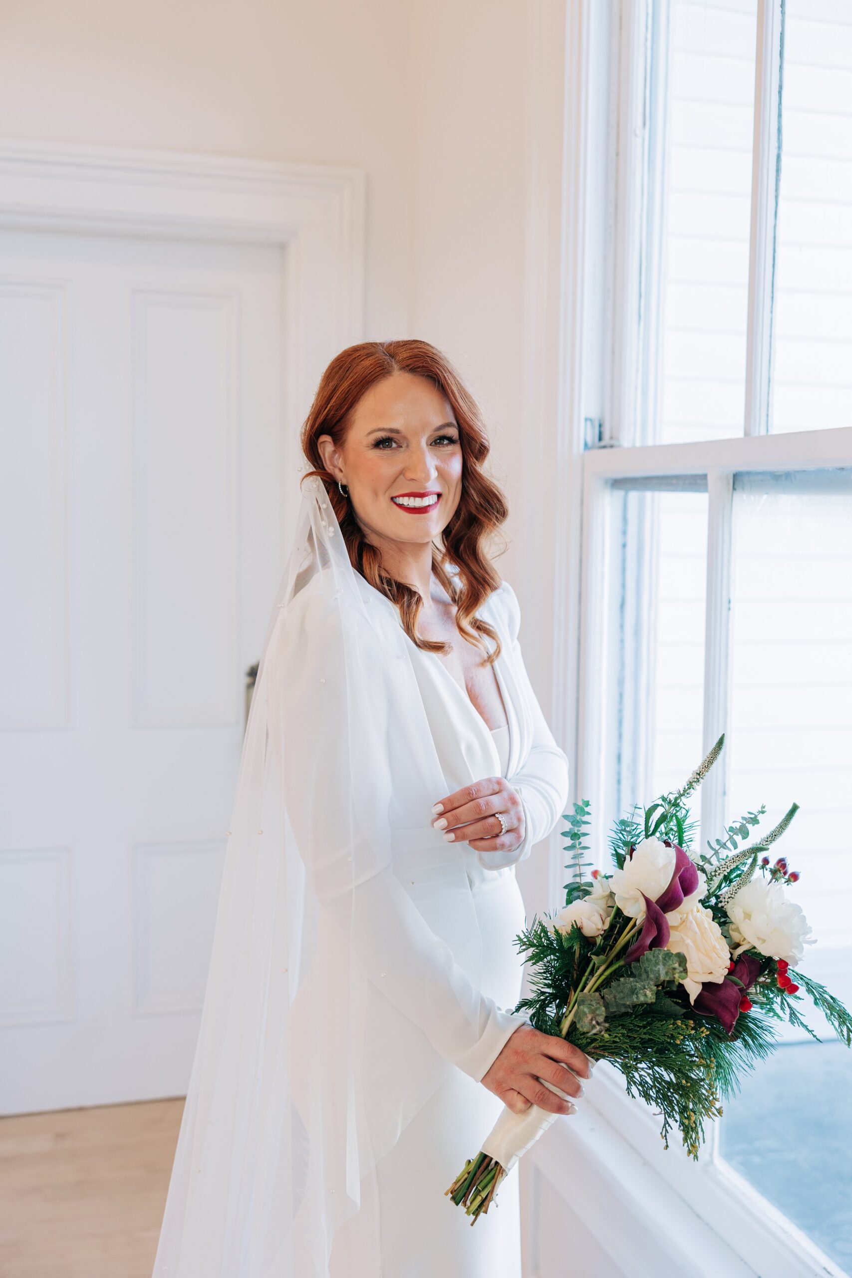 A bride smiles in her gown while standing in a window holding a red and white bouquet