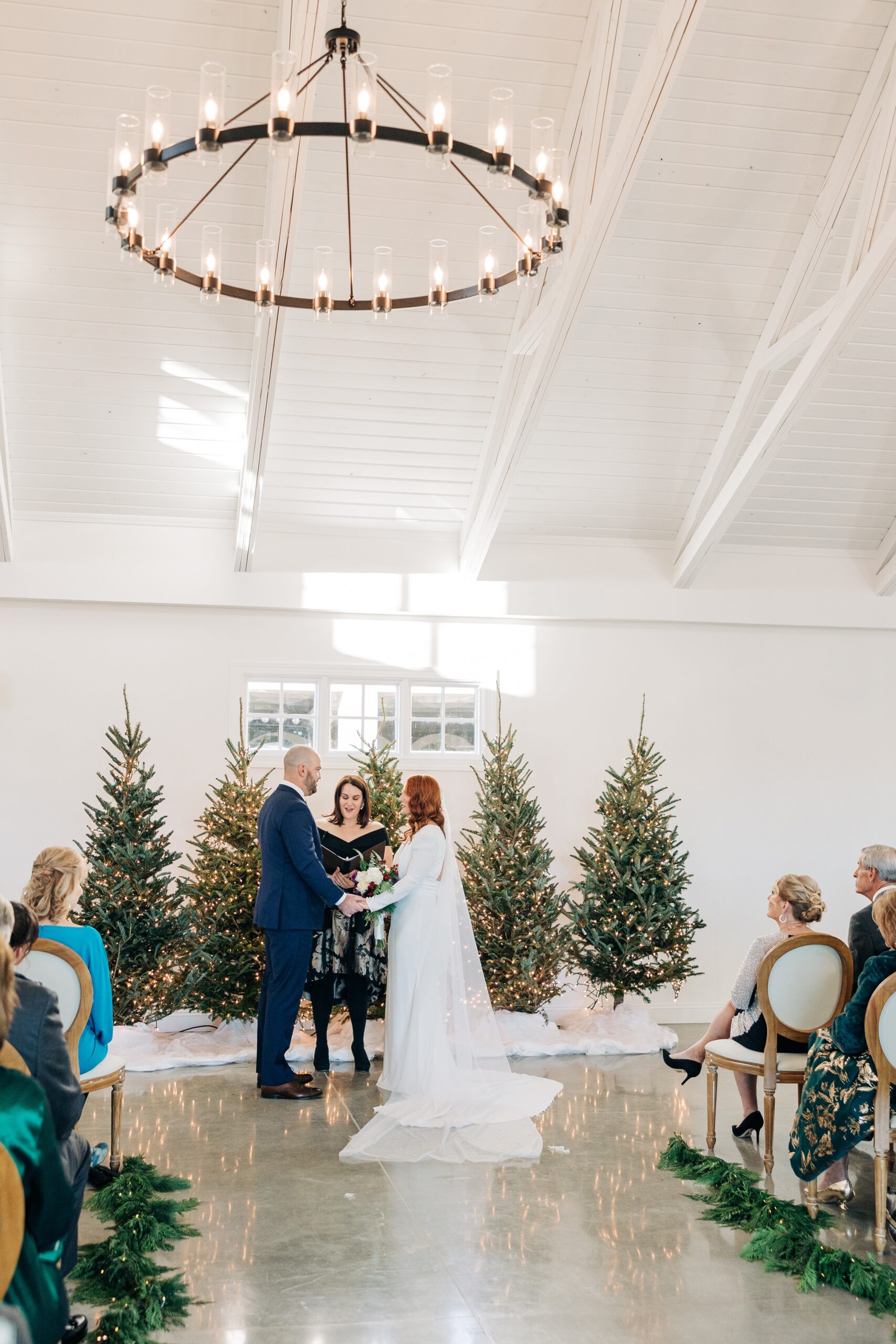 A bride and groom stand at the altar with christmas trees holding hands and listening to the officiant