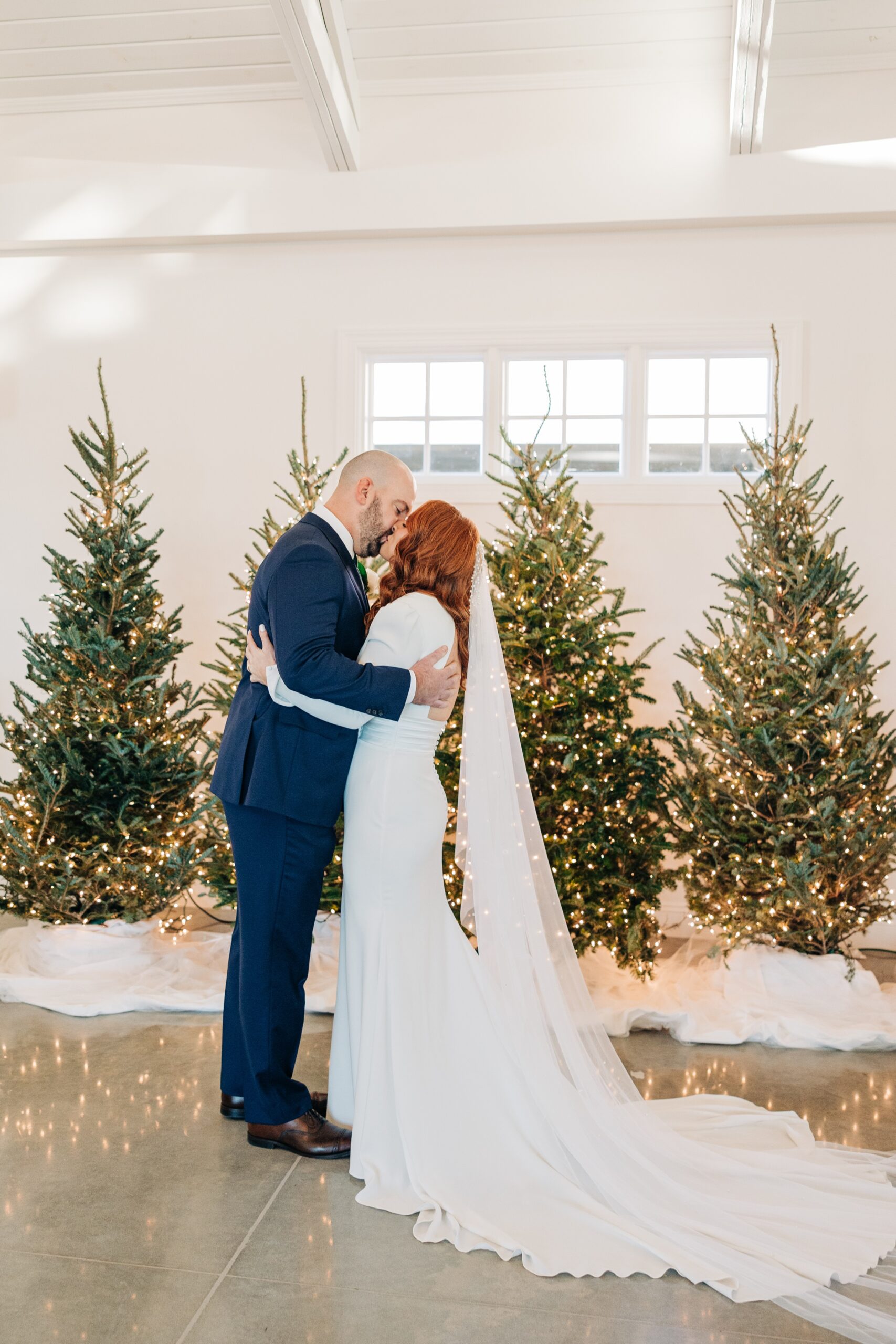 Newlyweds kiss by some christmas trees inside