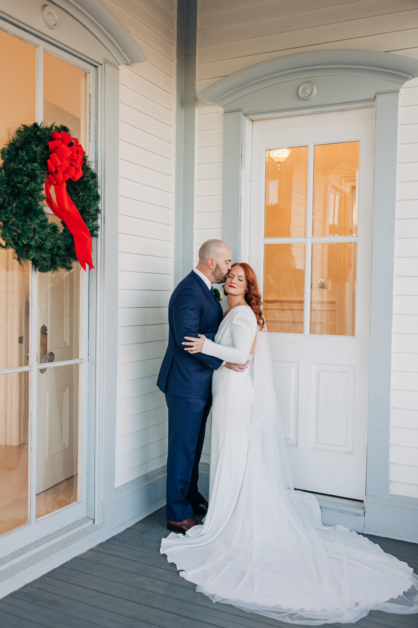 A groom in a blue suit kisses his bride on the temple next to a christmas wreath