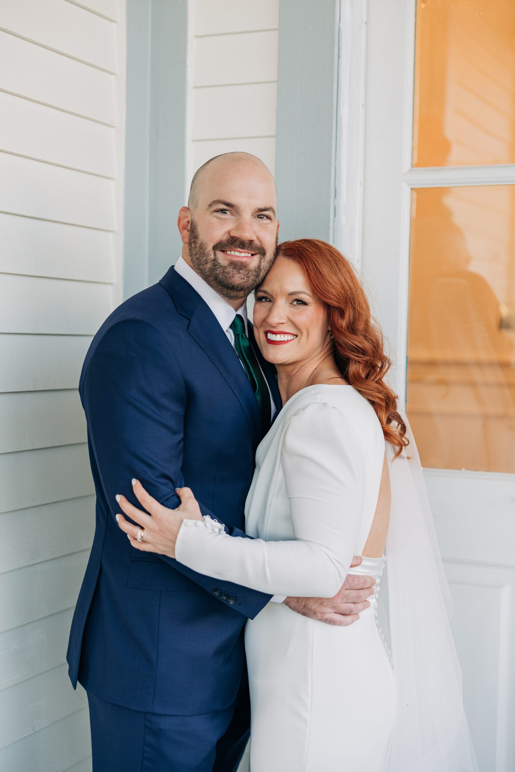 Newlyweds stand cheek to cheek on a porch with a blue suit and white gown