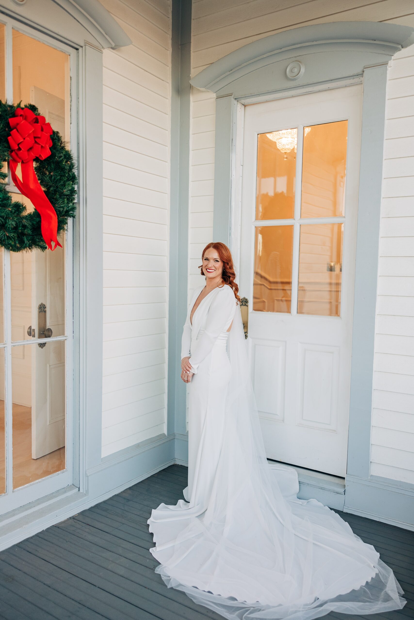 A bride stands on a porch with a christmas wreath