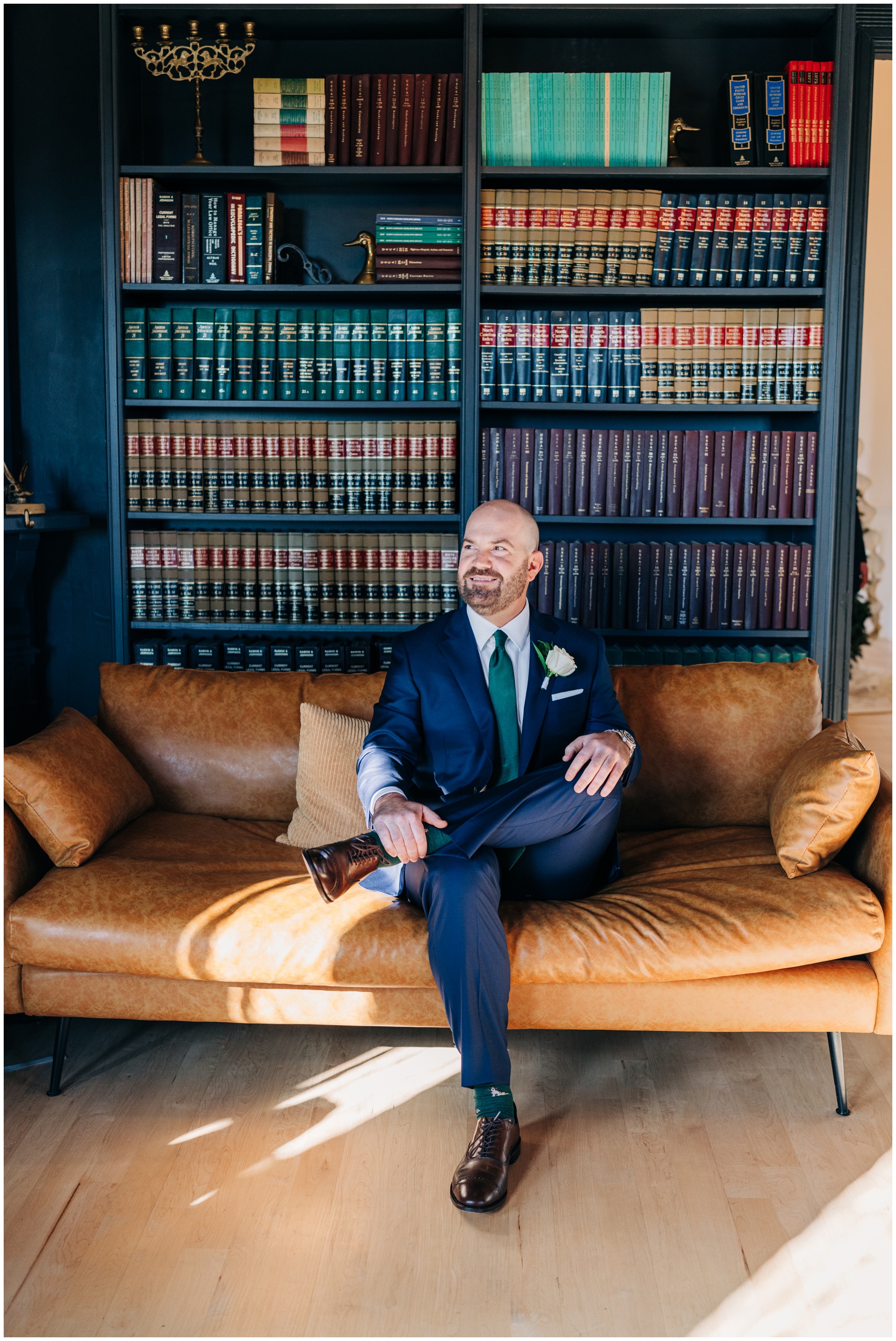 A smiling groom sits on a leather couch in a library in a blue suit