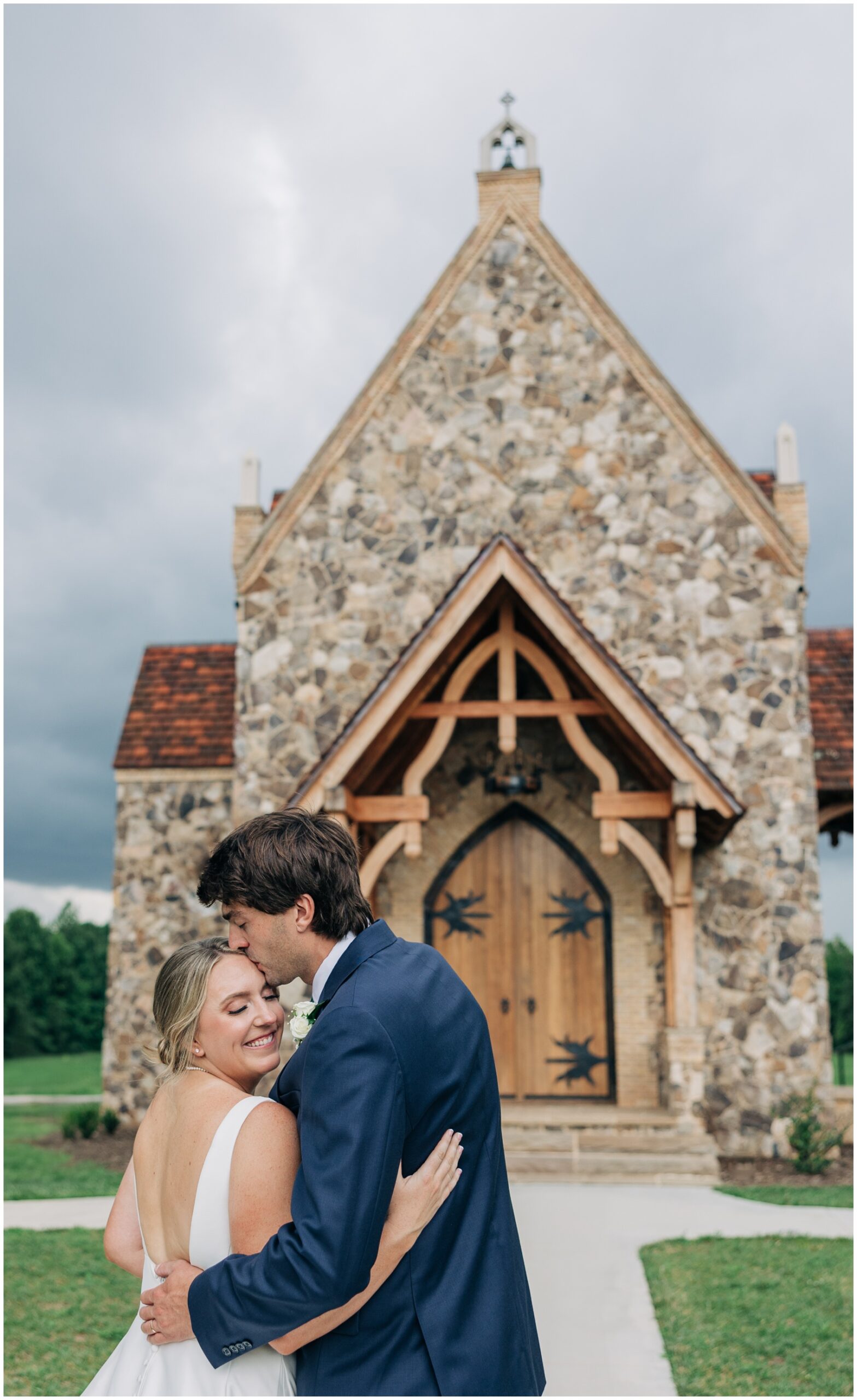 A groom kisses his smiling bride's head as they snuggle outside one of the Winston Salem Wedding Venues at the little chapel