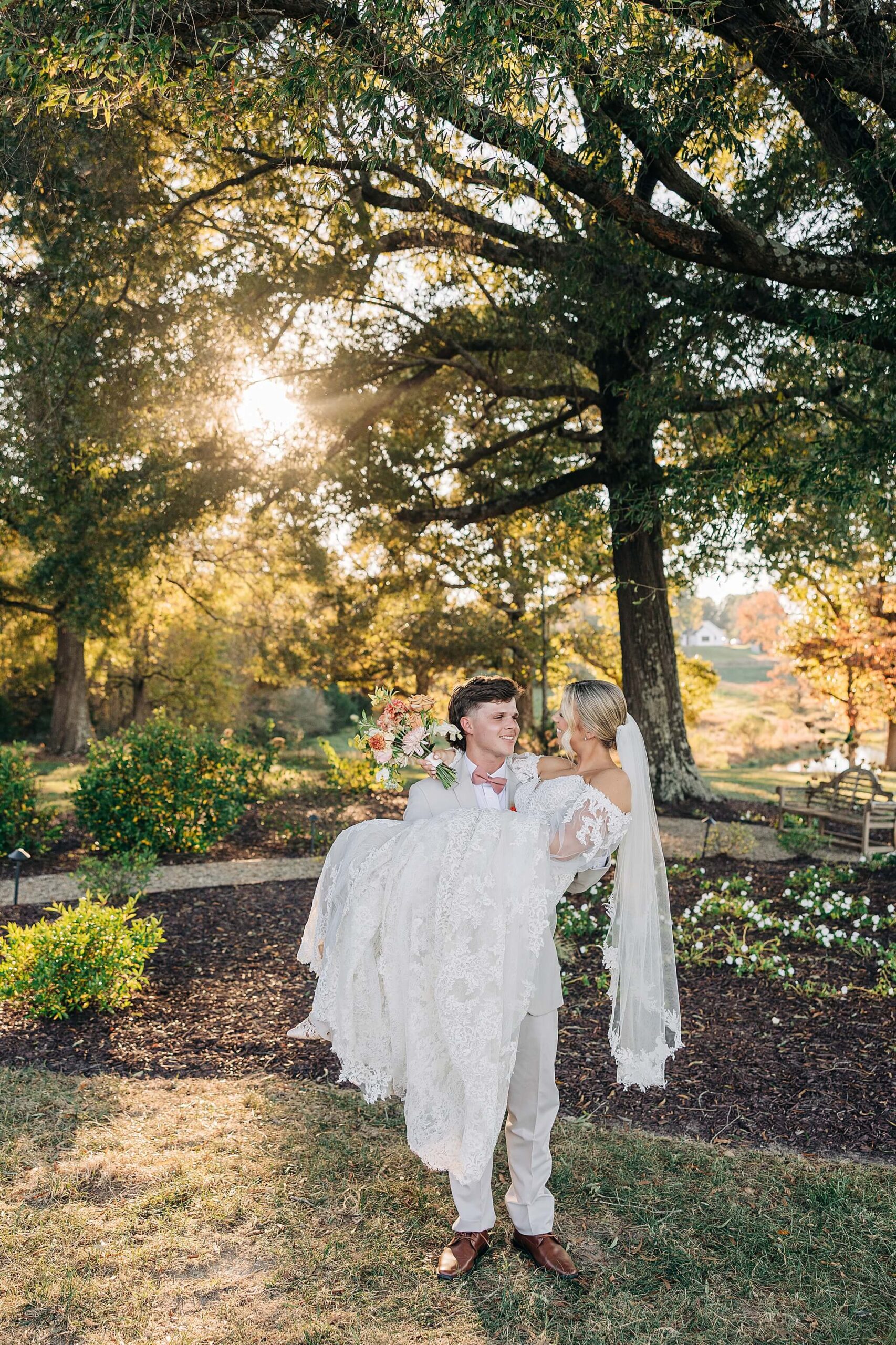 A groom carries his bride in her lace gown through a garden at sunset