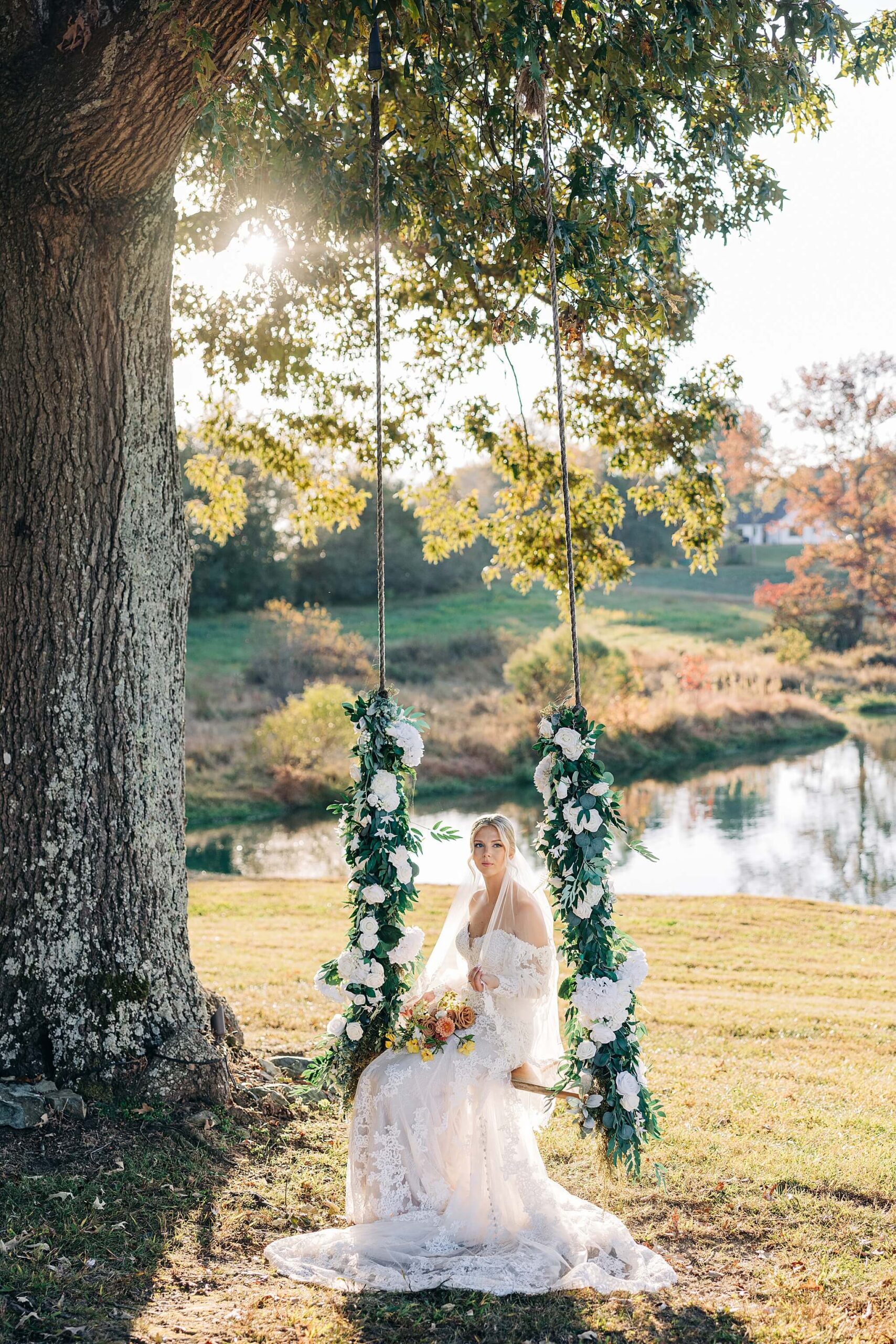 A bride in a lace gown sits on a flower covered tree swing holding her bouquet by a pond at Hayfield at Murchison Farms
