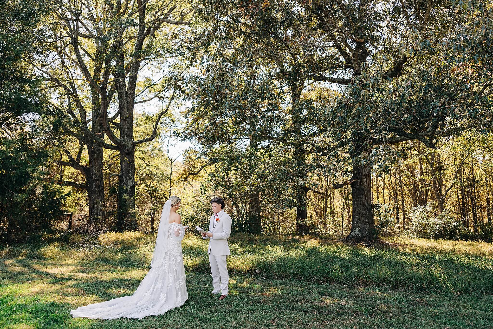 A groom reads a note to his bride alone on a grass lawn at sunset near a forest