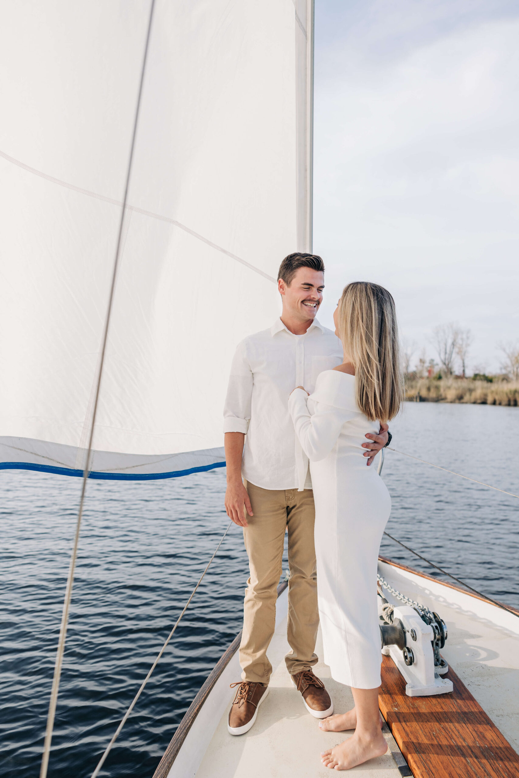 A man laughs while cuddling with his fiancee on the bow of a sailboat at sunset during engagement photos in wilmington nc