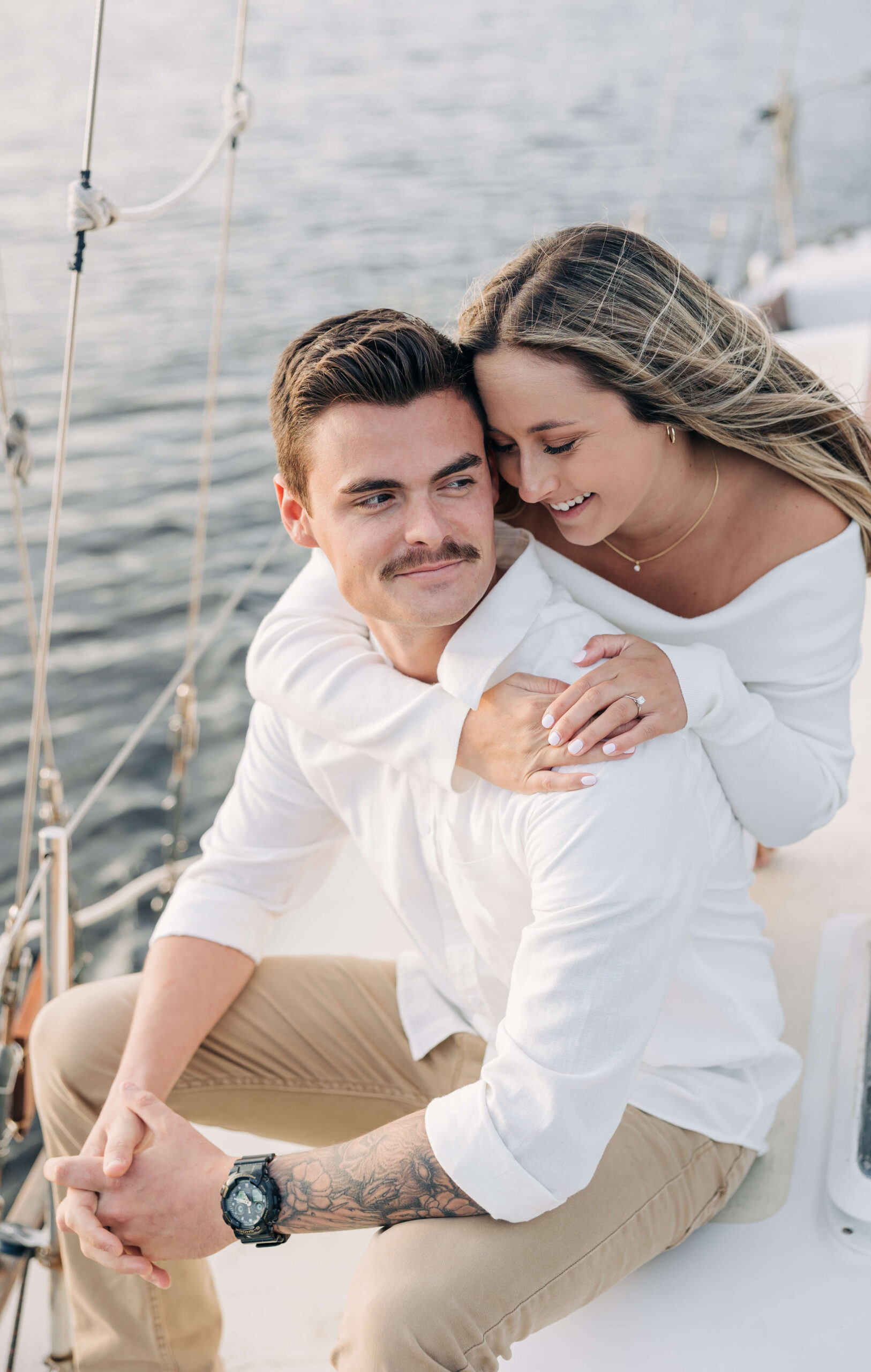 A woman in a white dress hugs onto her fiancee as they enjoy a sail at sunset