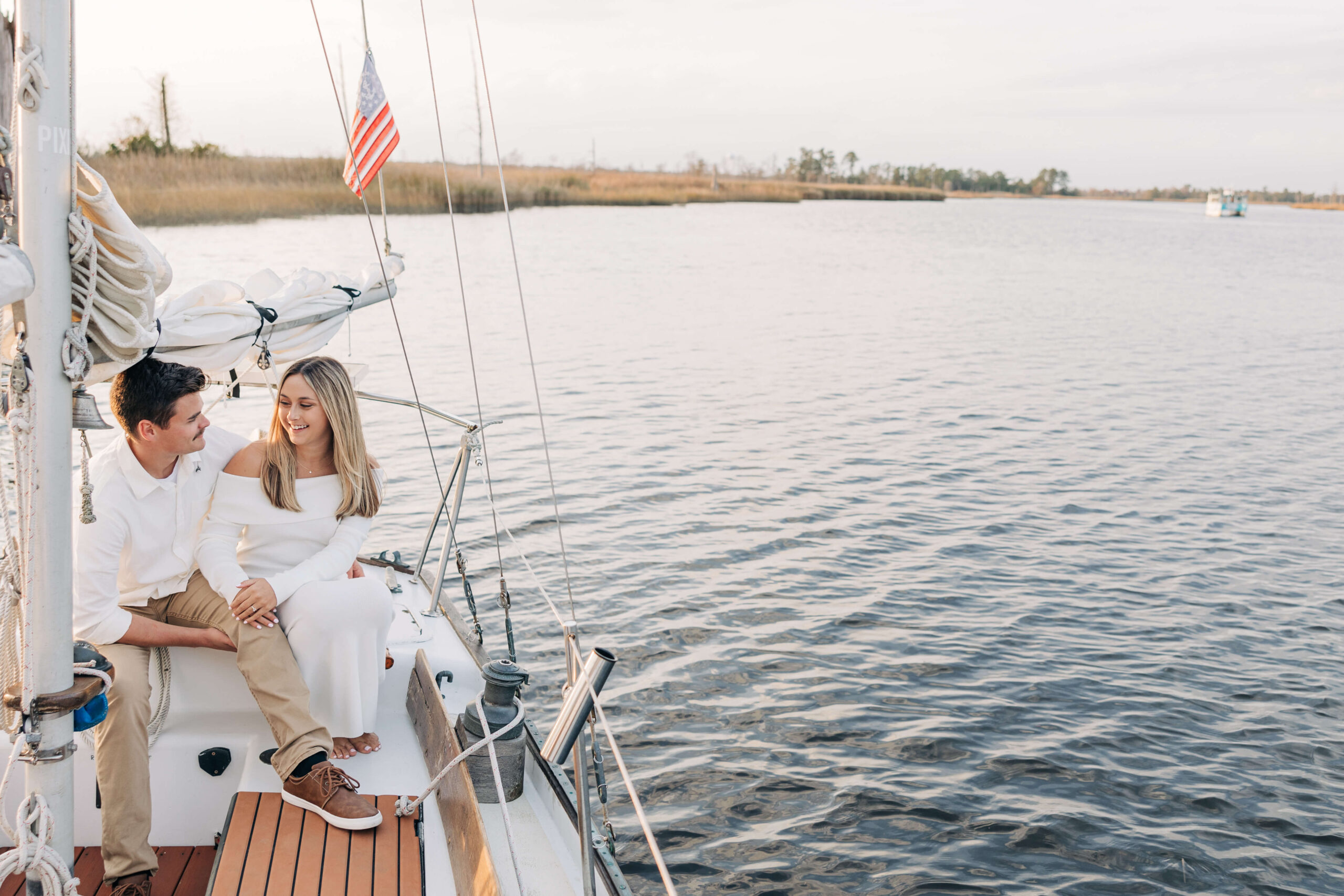 Newly engaged couple dressed in white sit on a sailboat at sunset laughing during their engagement photos in wilmington nc