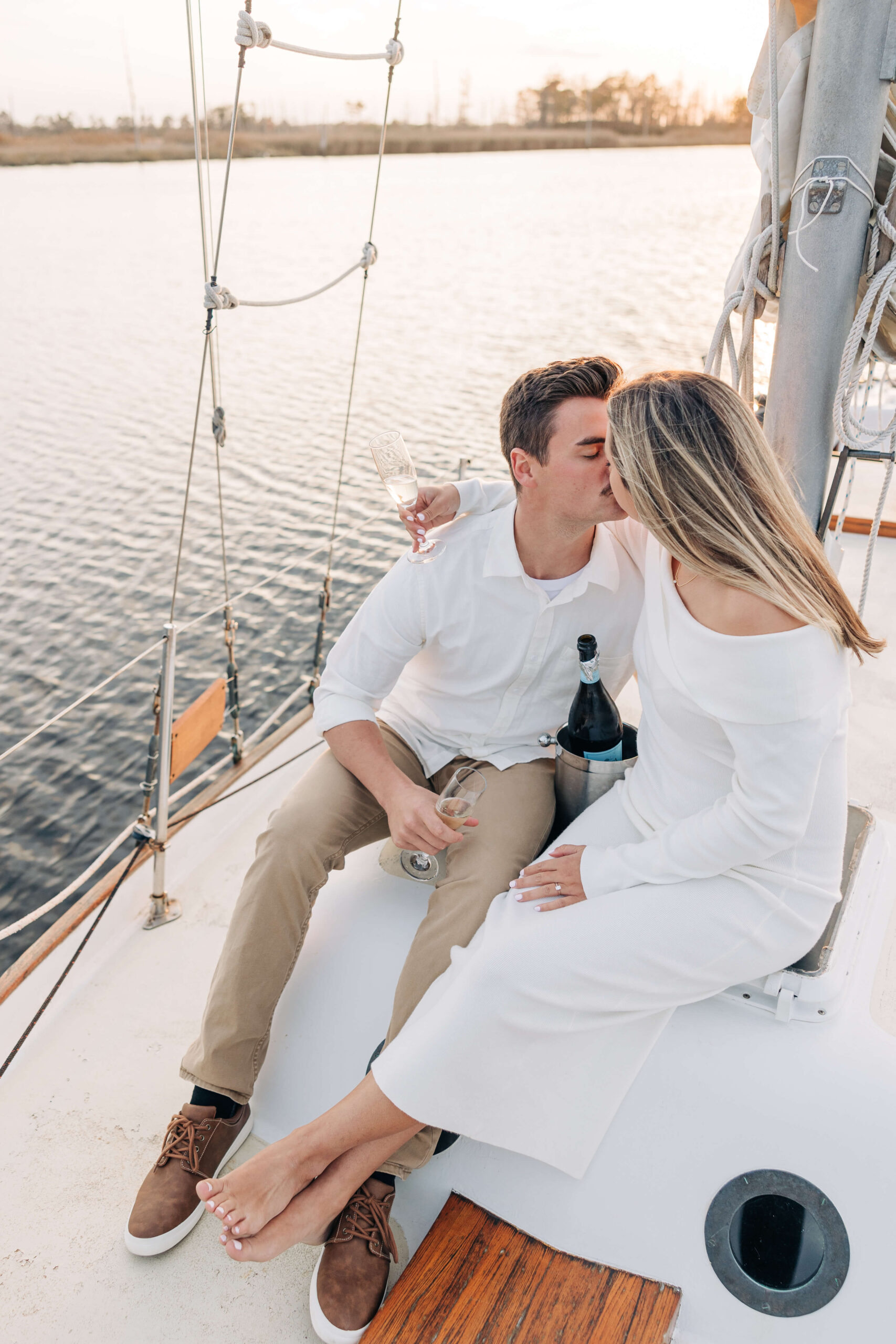 A happy newly engaged couple share a kiss while holding champagne on a sailboat at sunset dressed in white during their engagement photos in wilmington nc