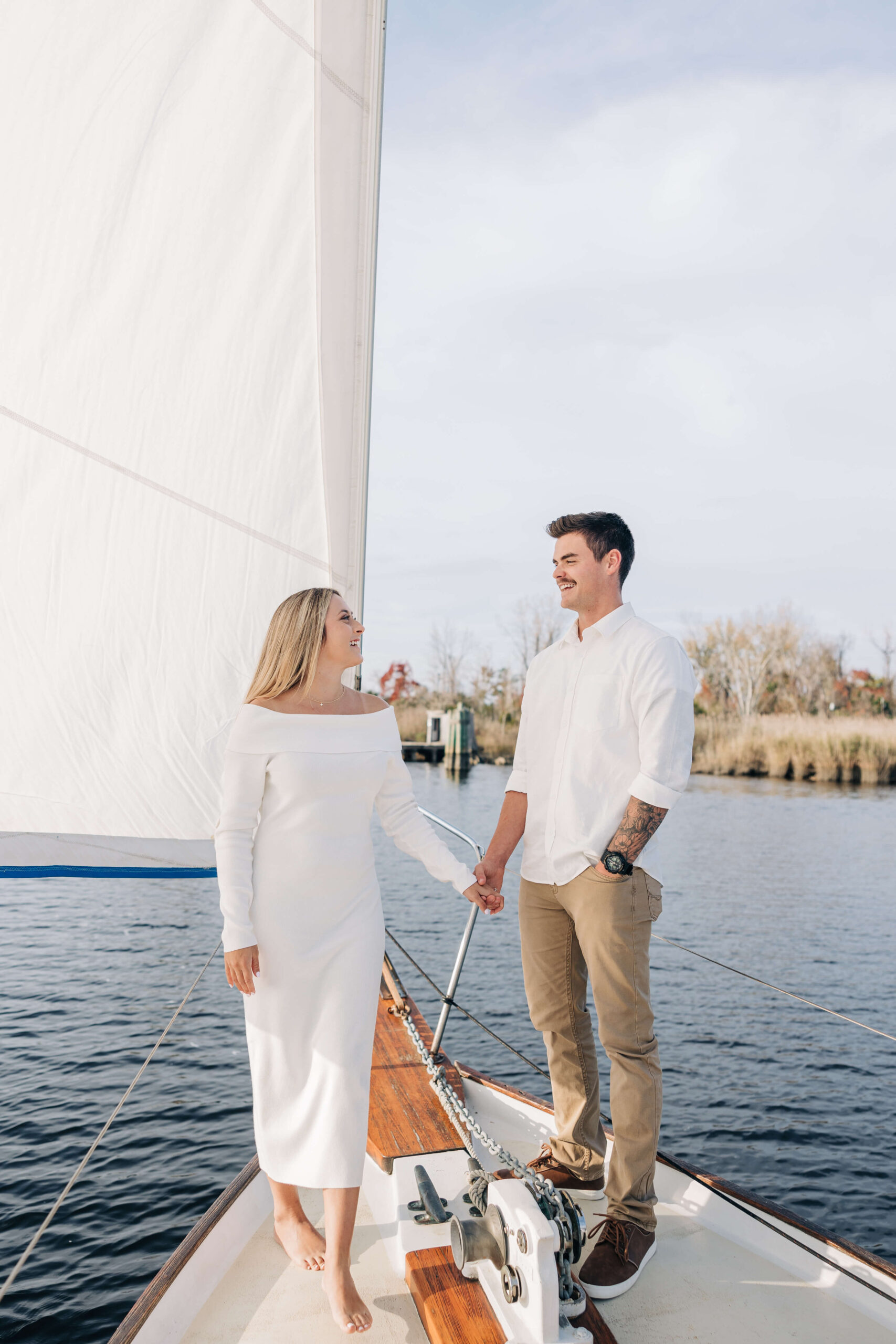 A happy couple laughs while holding hands and standing on the bow of a sailboat at sunset during engagement photos in wilmington nc