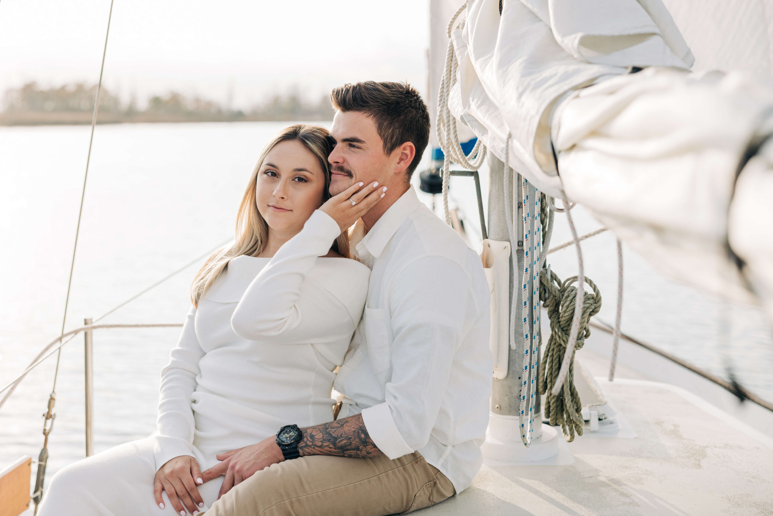 A woman sits in the lap of her fiancee in a white dress with a hand on his chin during engagement photos in wilmington nc