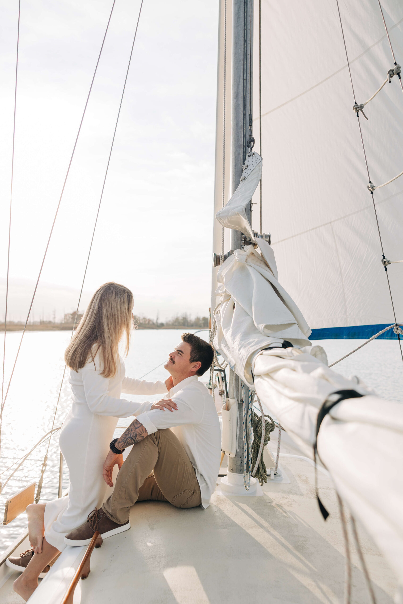 A happy couple cuddle on a sailboat at sunset during their engagement photos in wilmington nc dressed in white