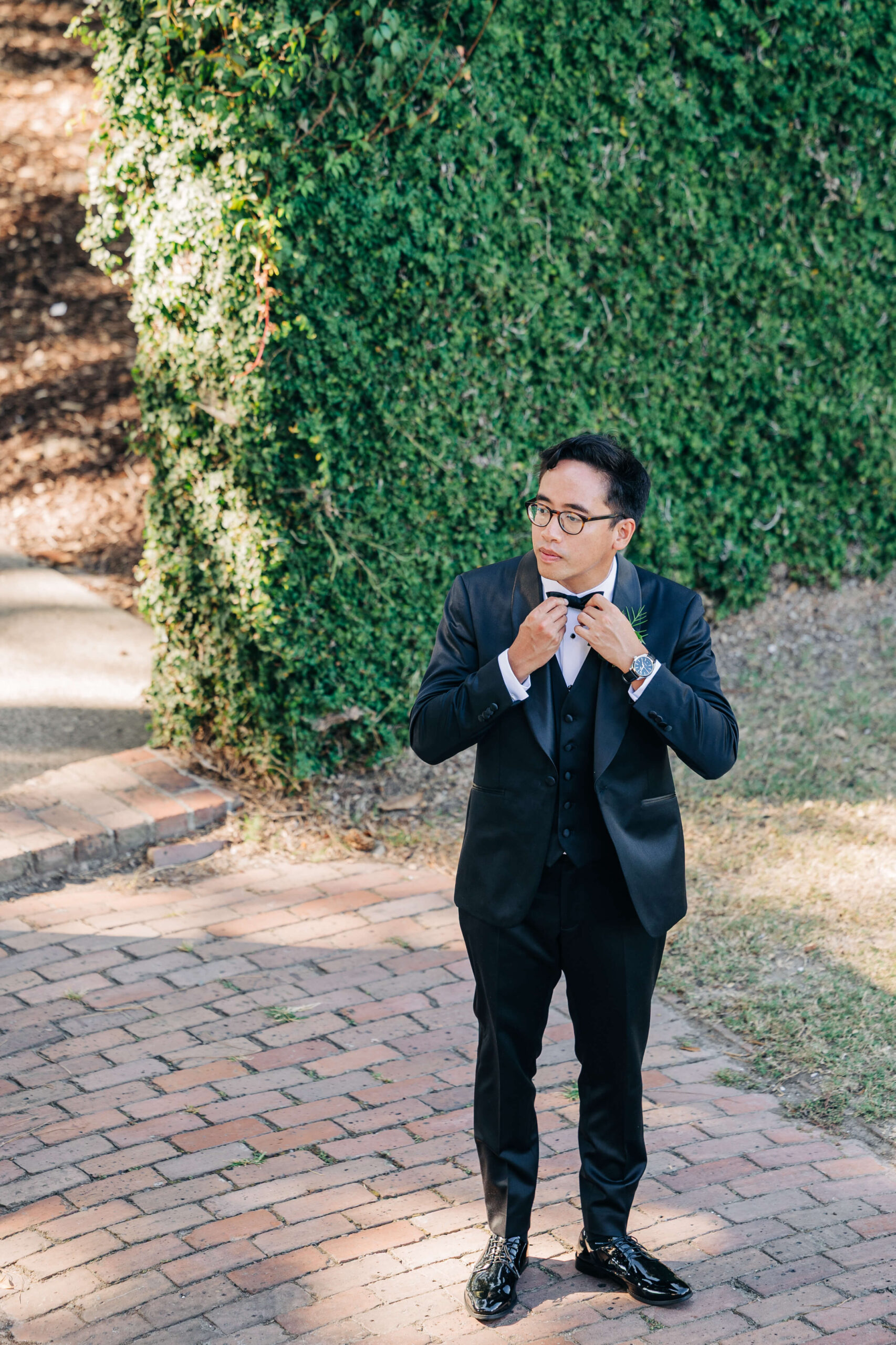 A groom adjusts his tie in a garden brick walkway