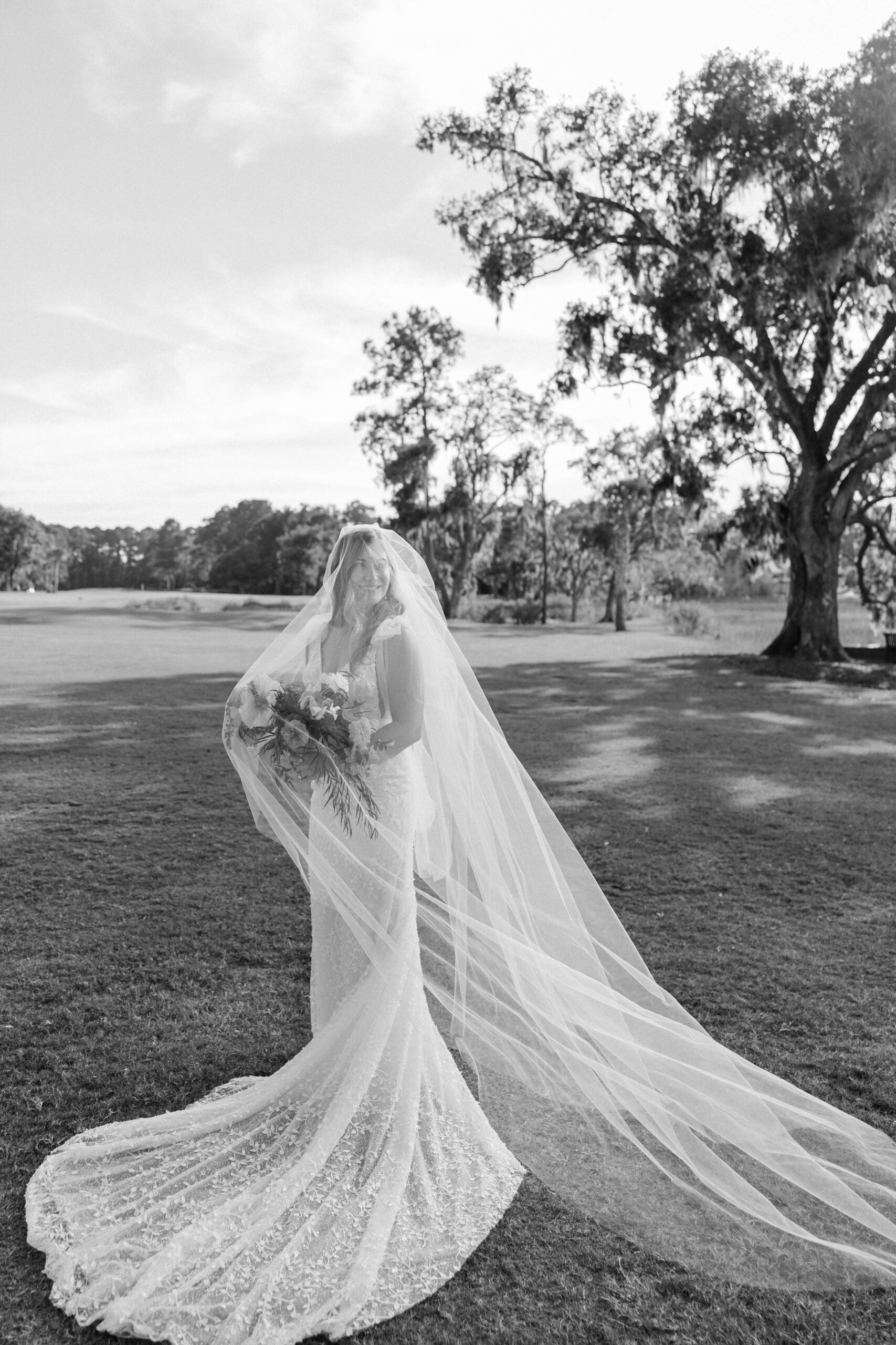 A happy bride smiles from under the veil in the Dunes West Golf Club wedding venue lawn while holding her bouquet
