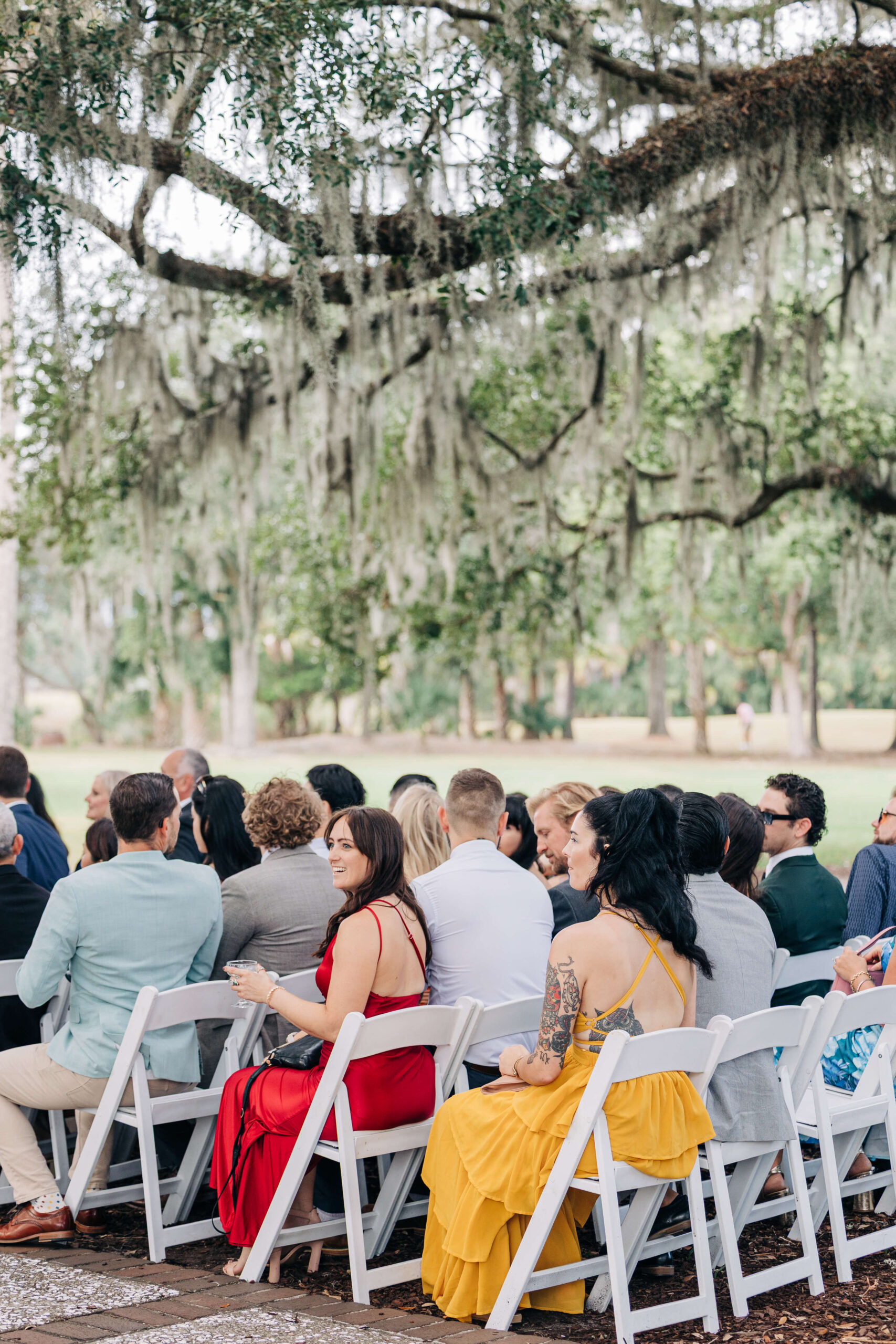 Wedding ceremony guests wait in white chairs under oak trees