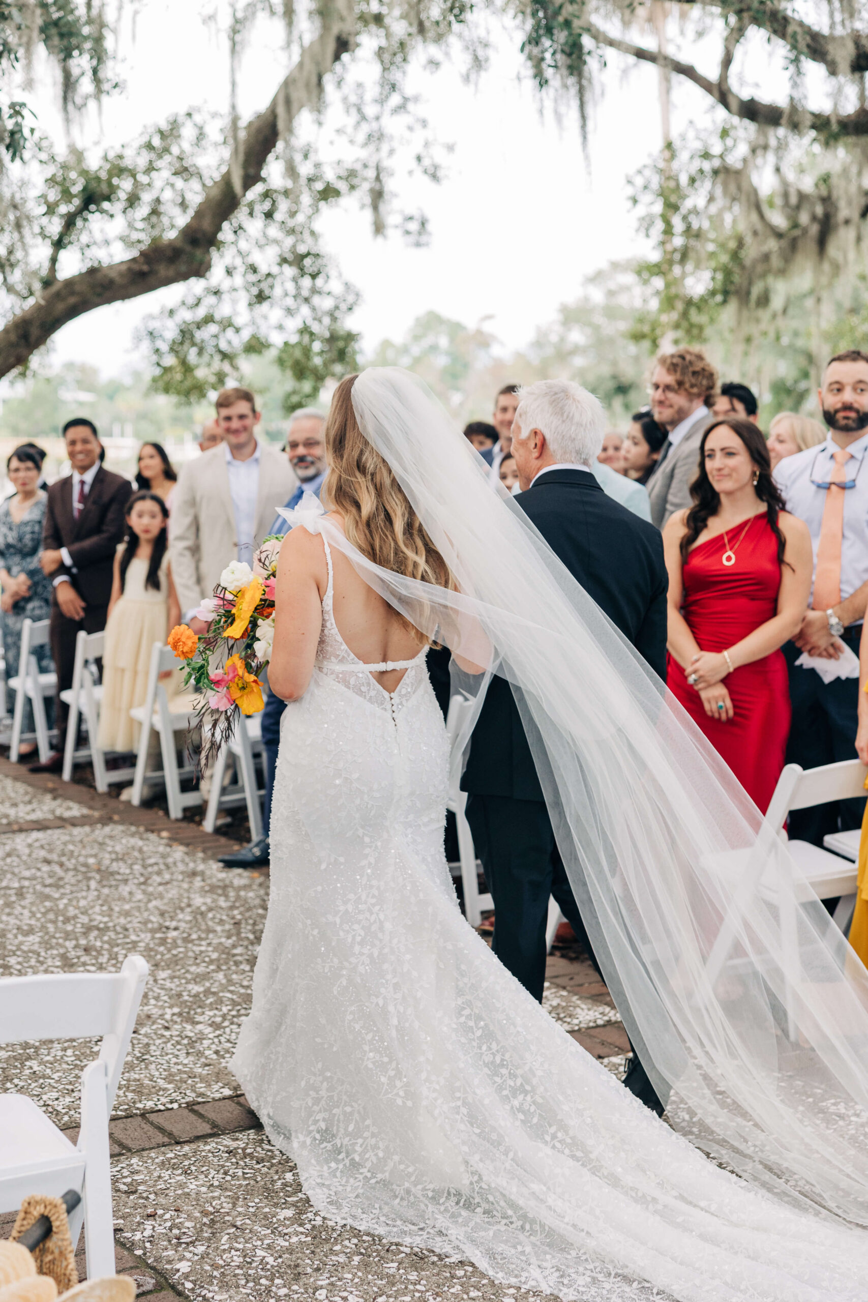 A bride walks down the aisle with dad wearing a long flowing veil as guests watch