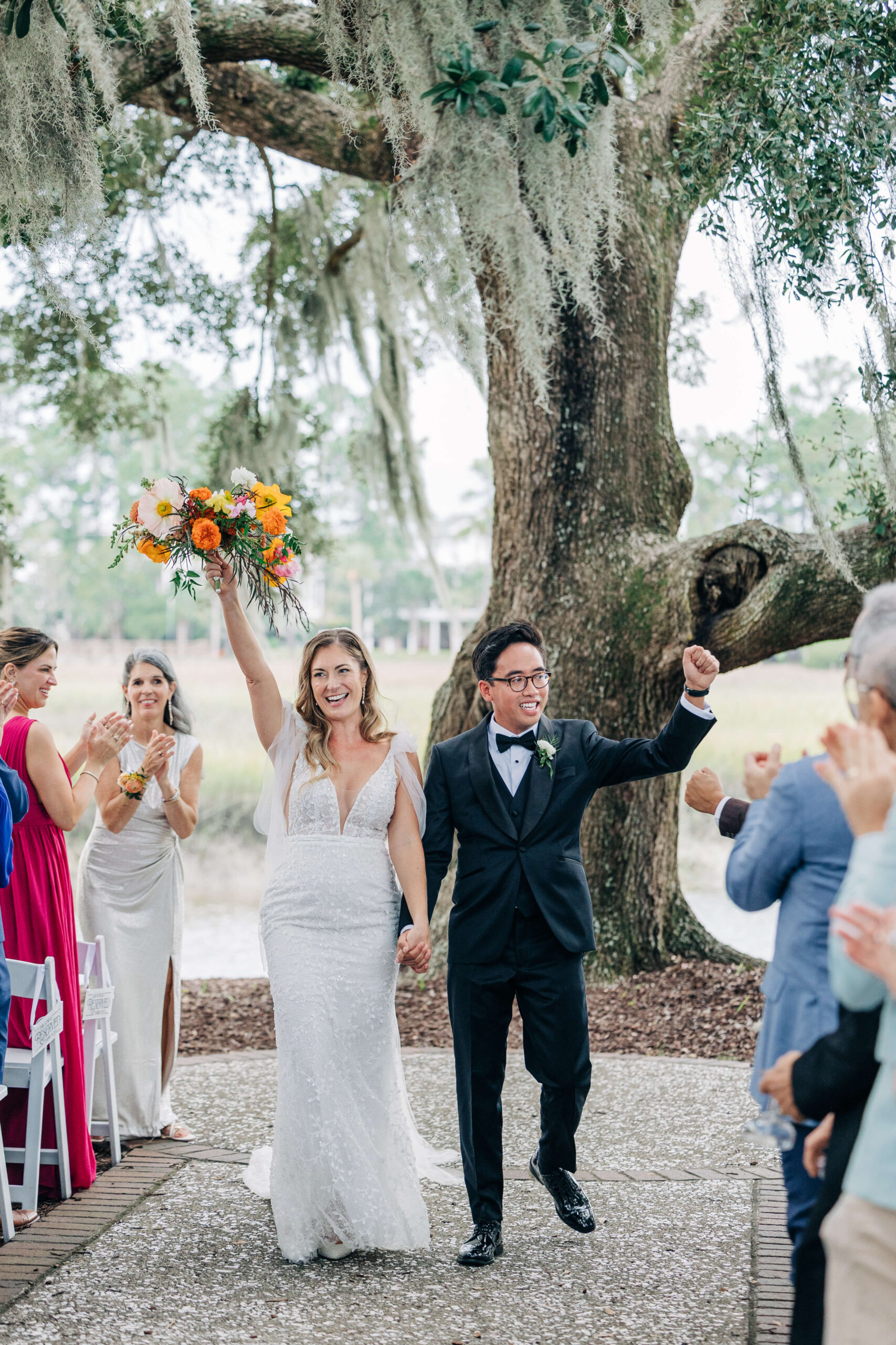 Newlyweds lift their arms to celebrate while exiting their Dunes West Golf Club wedding ceremony outdoors under oak trees