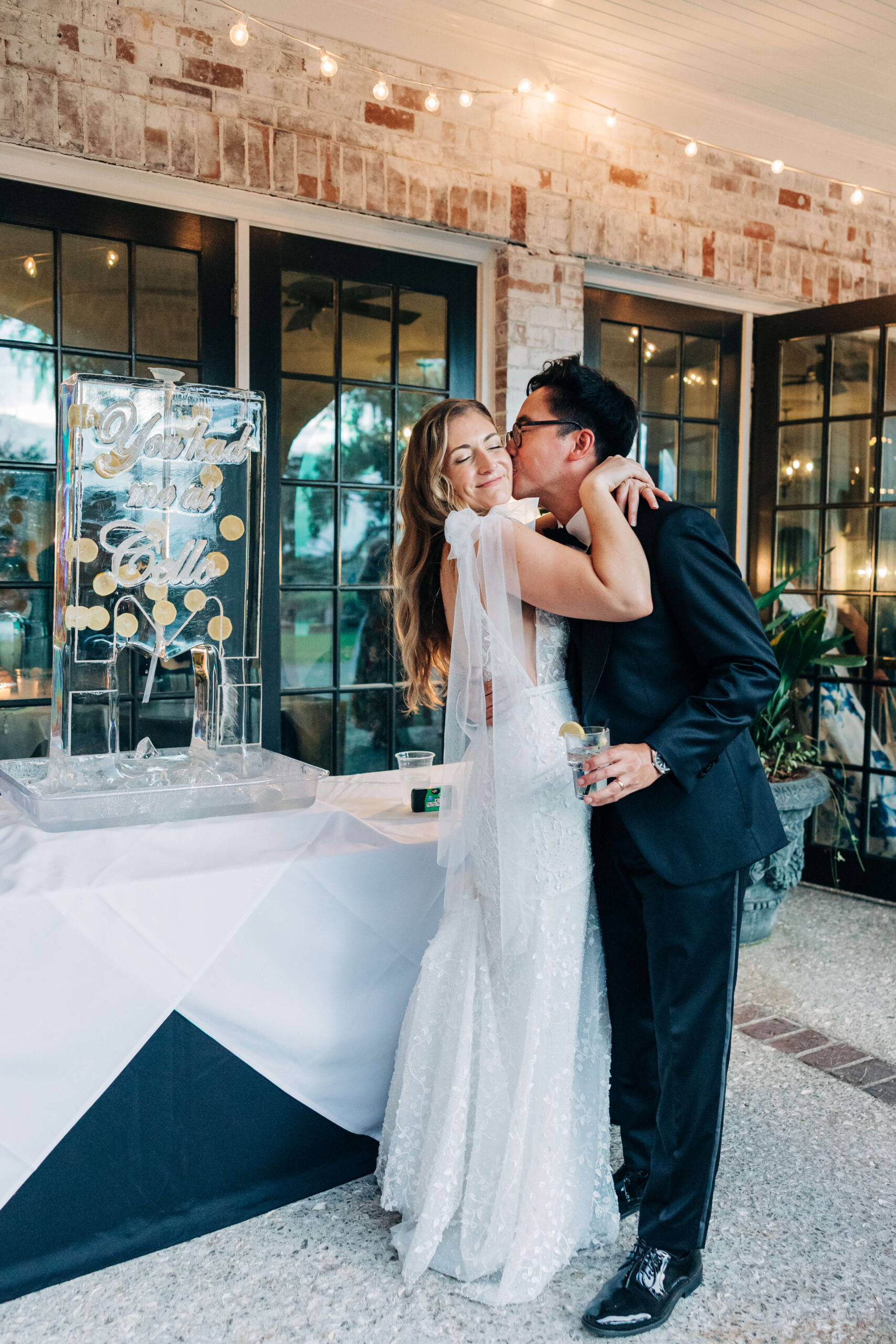 A smiling bride is kissed on the cheek by her groom by a custom ice sculpture during their Dunes West Golf Club wedding