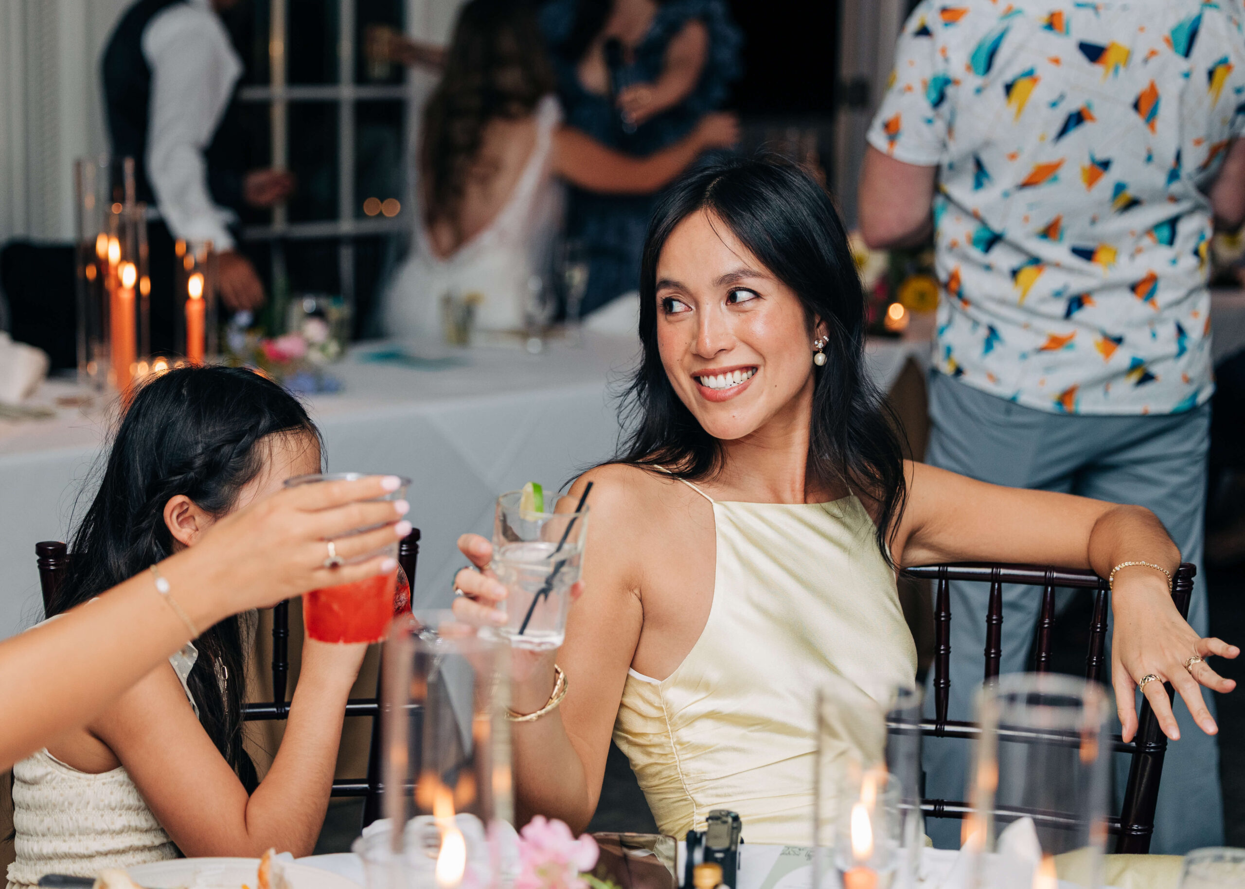 A happy woman in gold leans back to toast with reception guests at their table