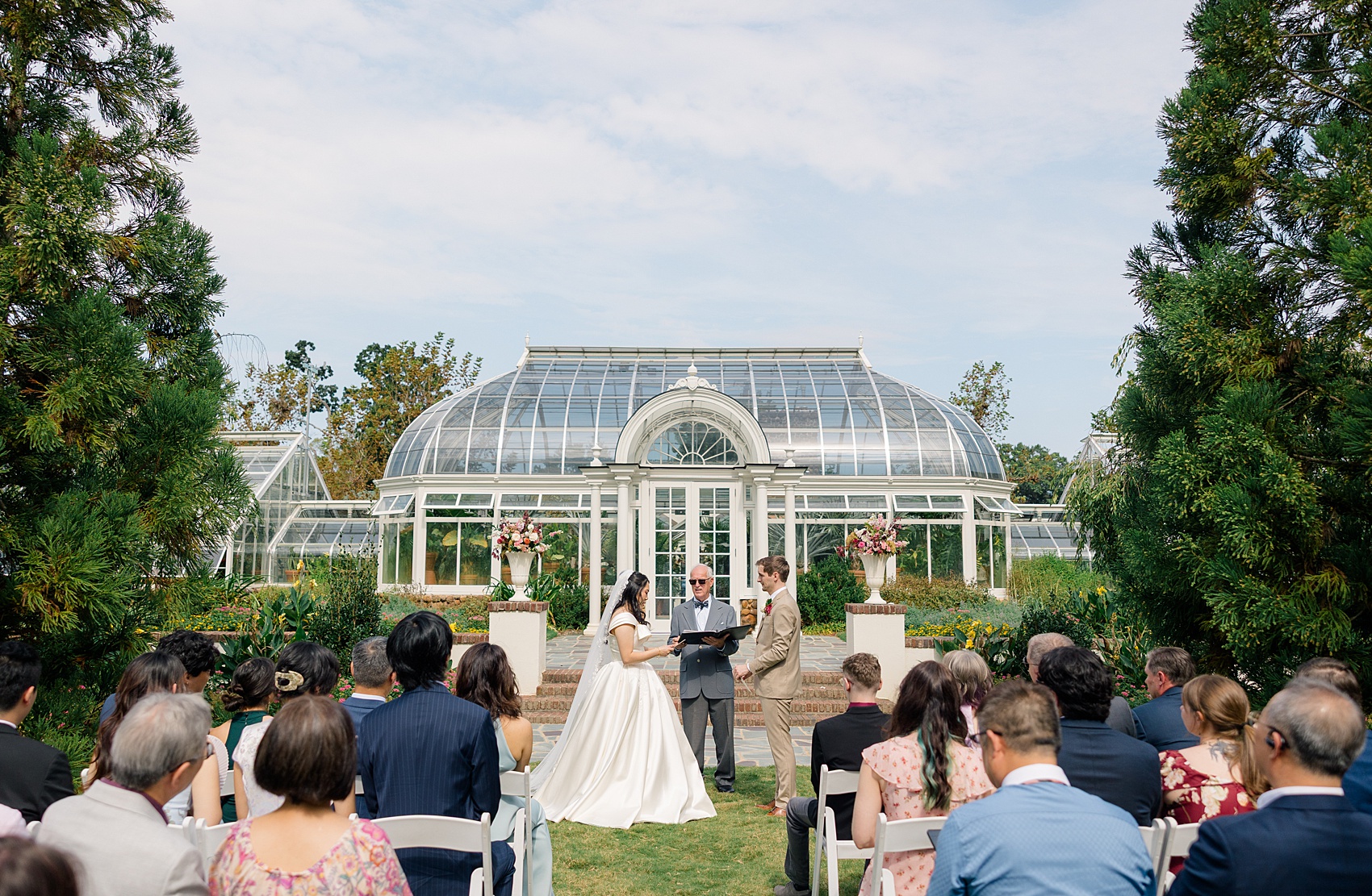 A bride and groom stand listening to the officiant during their outdoor garden ceremony at the barn at reynolda village wedding venue