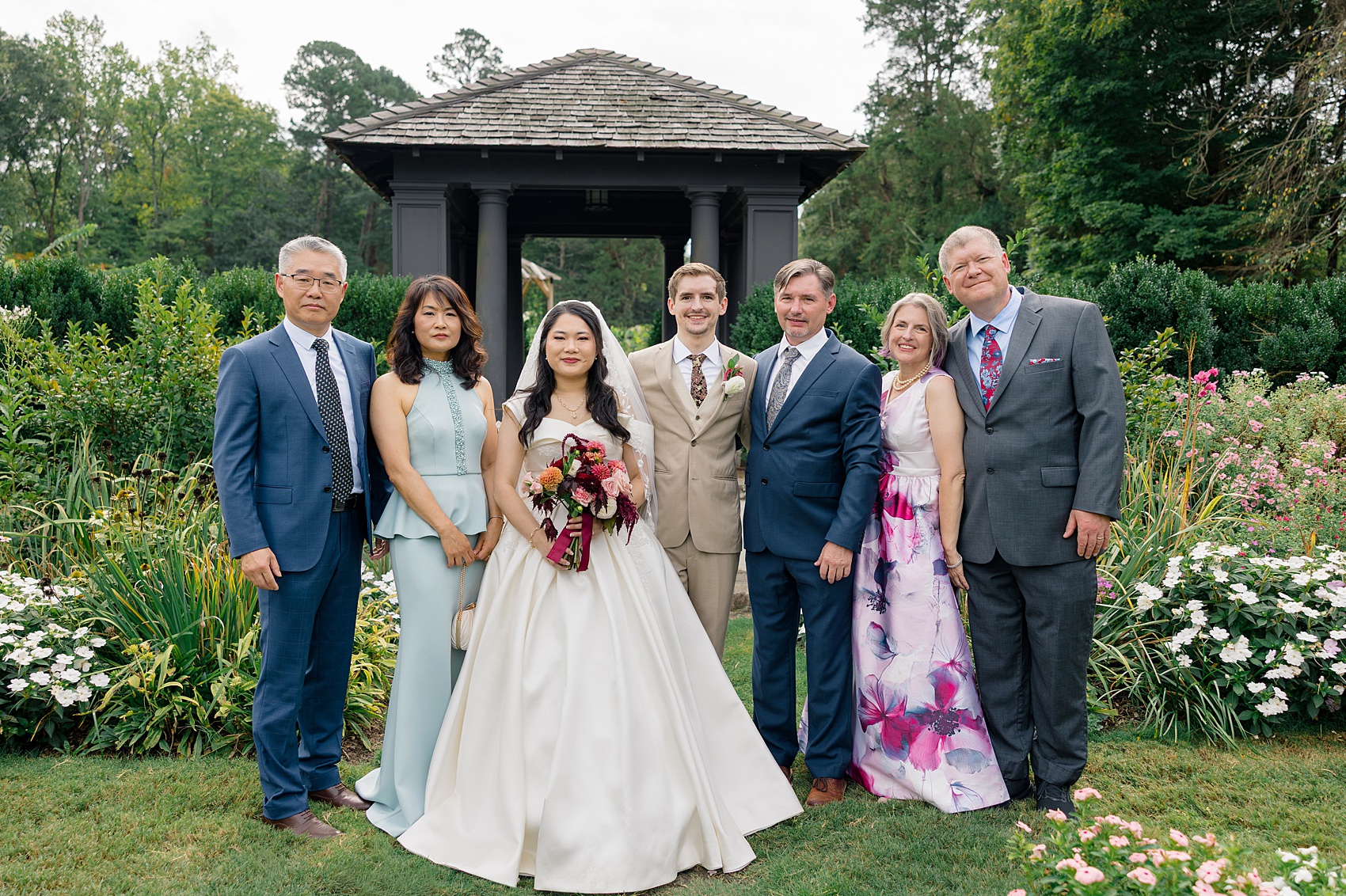 Newlyweds smile while standing with their parents in a garden