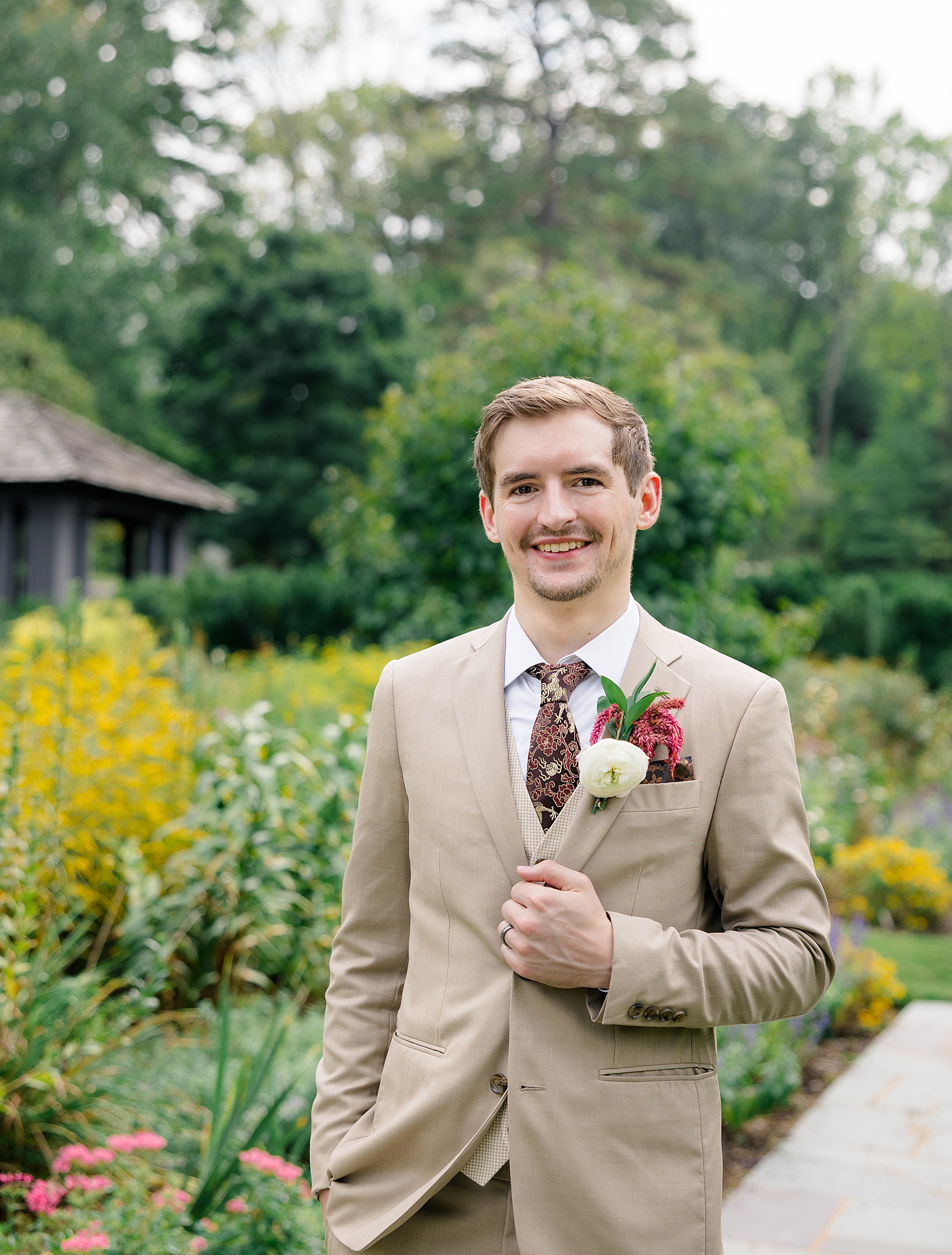 A happy groom smiles in a tan suit holding his lapel in a garden